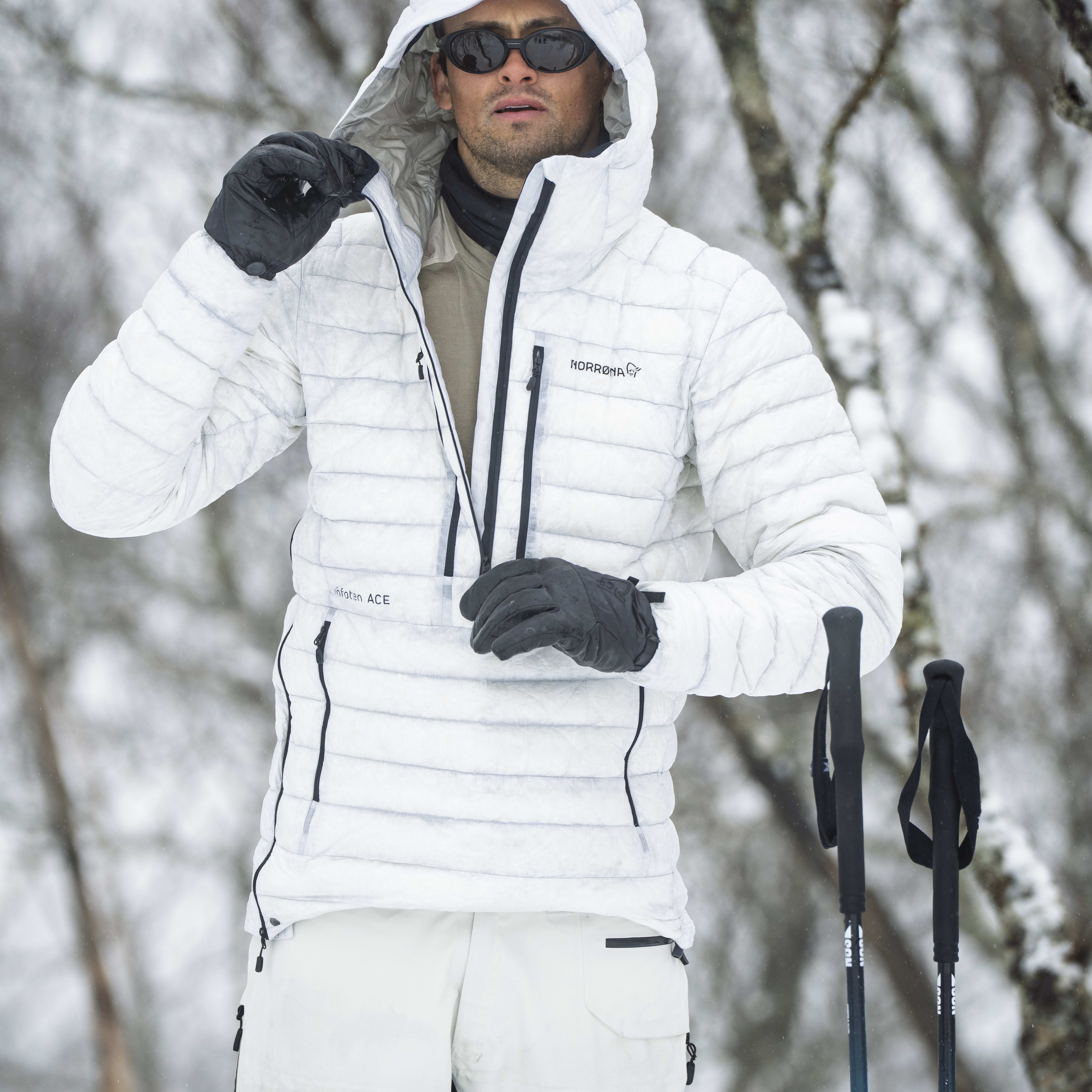 A man wearing a white Norrøna jacket and pants stands in a snowy forest holding trekking poles.