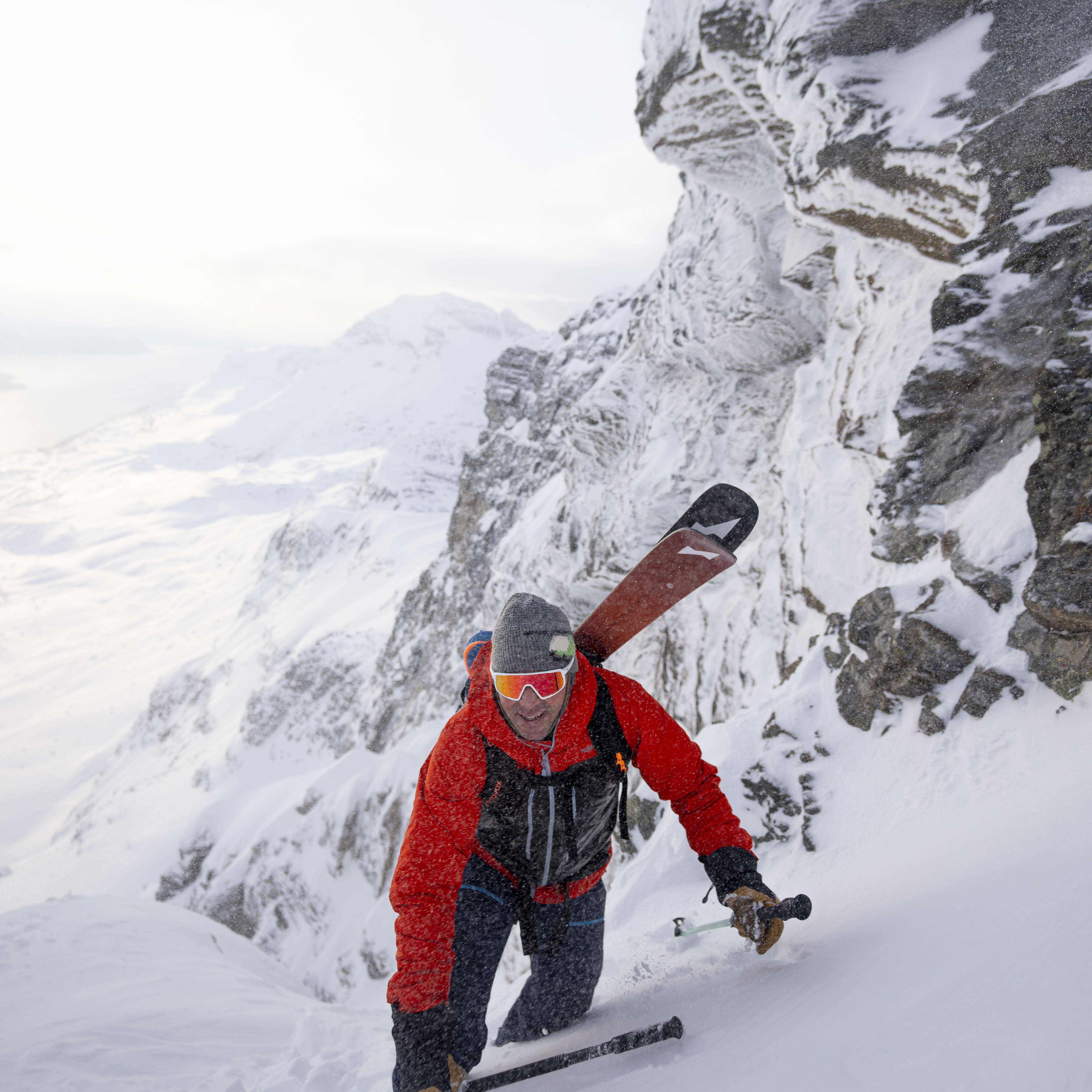 A skier with skis on their back, climbs a snow-covered mountain. They wear a red Norrøna jacket, goggles, and a hat.
