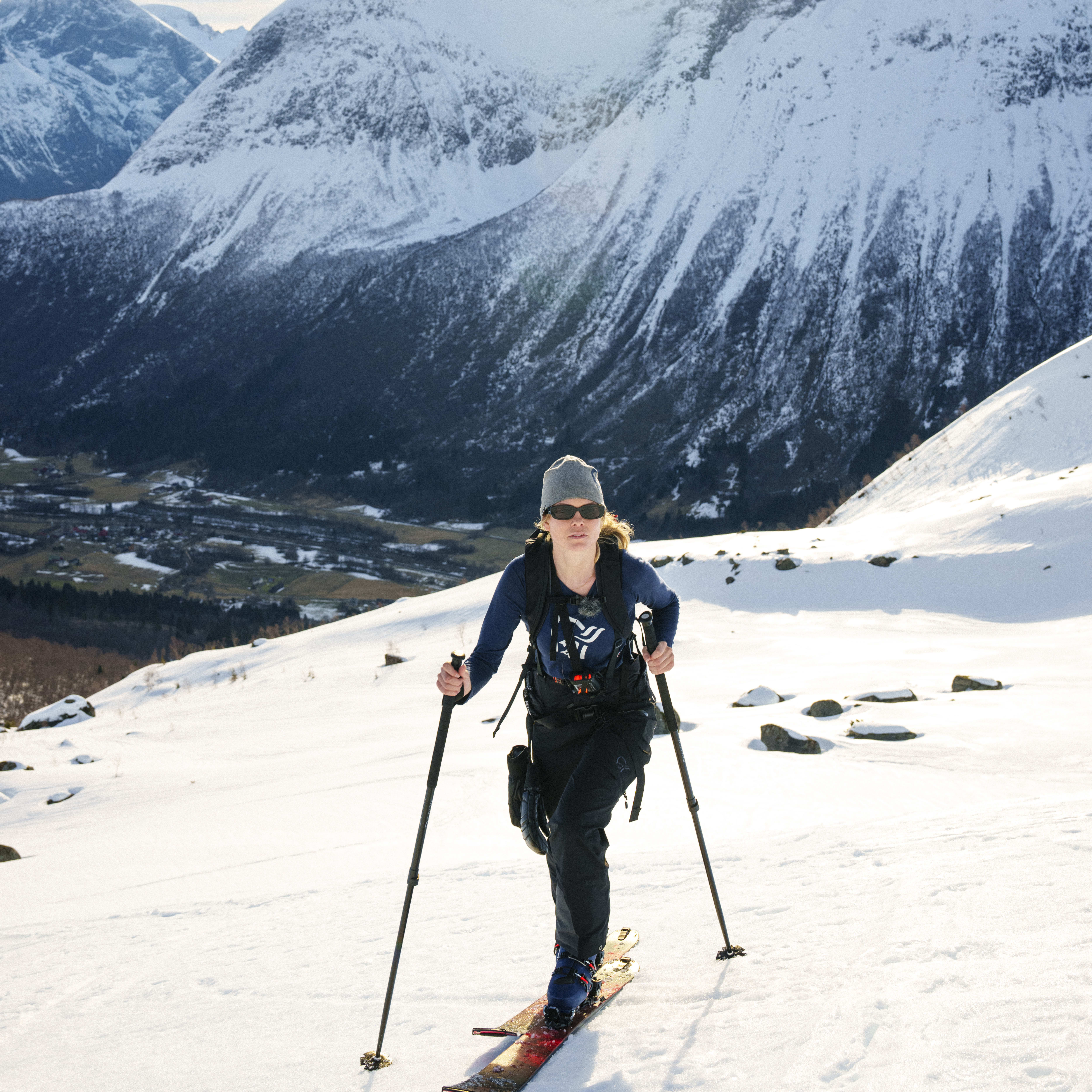 A woman ski touring on snow with mountain backdrop. She wears a hat, sunglasses and Norrøna pants.
