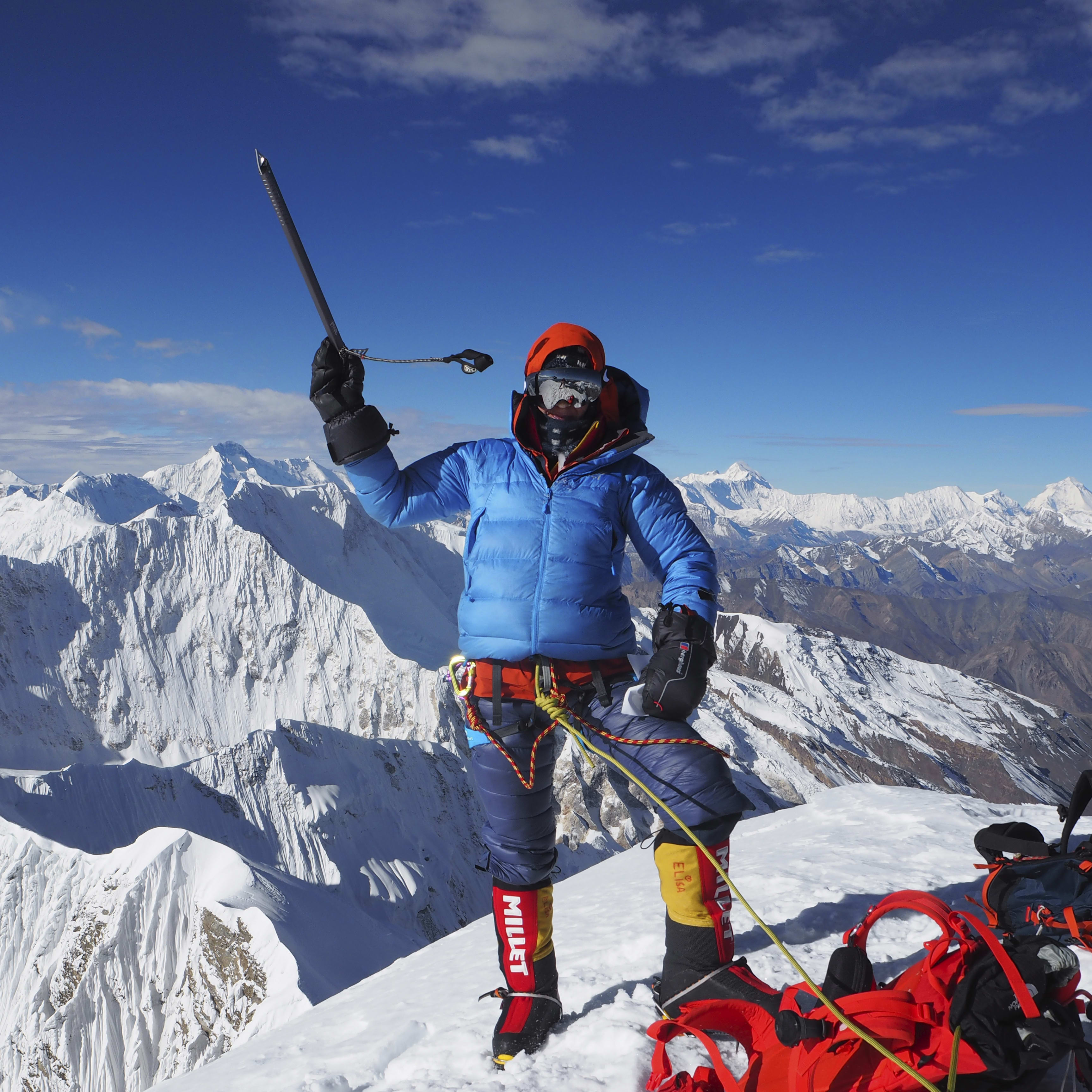 A climber stands on a snowy mountain peak, wearing a Norrøna jacket and holding an ice ax. The climber is silhouetted against a clear blue sky with a few puffy clouds. The view of the snow-capped mountains is breathtaking.