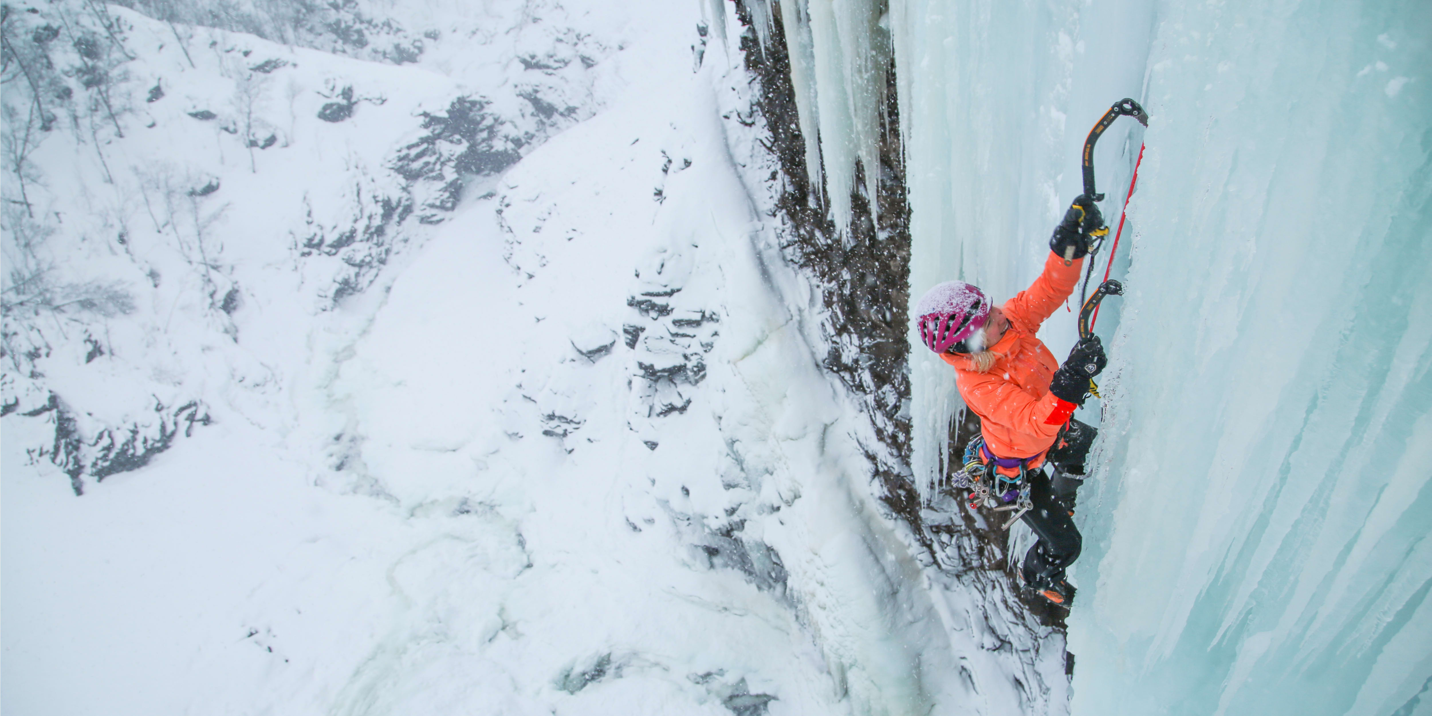 A climber wearing a Norrøna jacket and helmet ascends an ice wall.  
