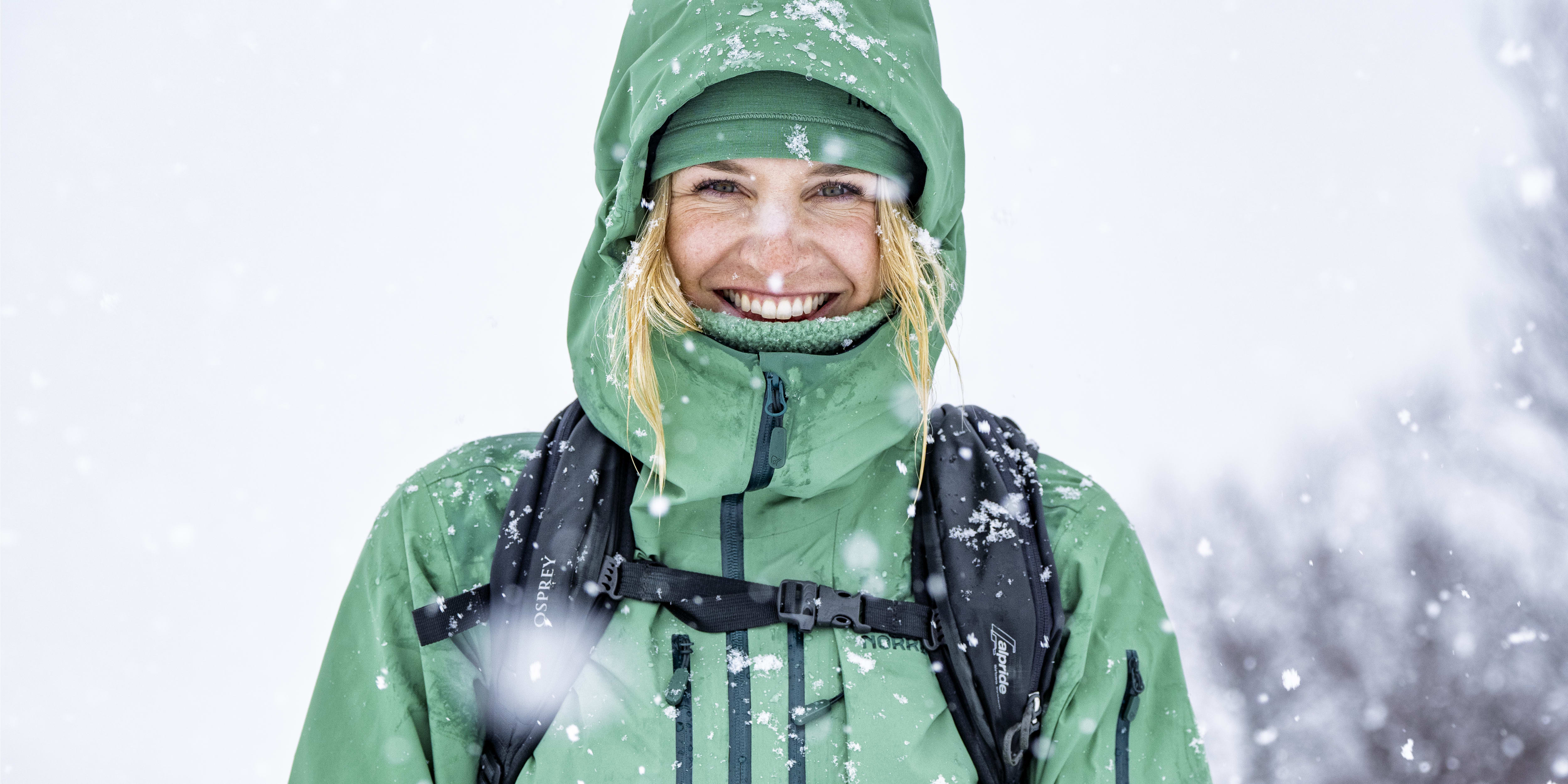 Smiling woman in a green Norrøna jacket with hood up, and black backpack, stands in falling snow.