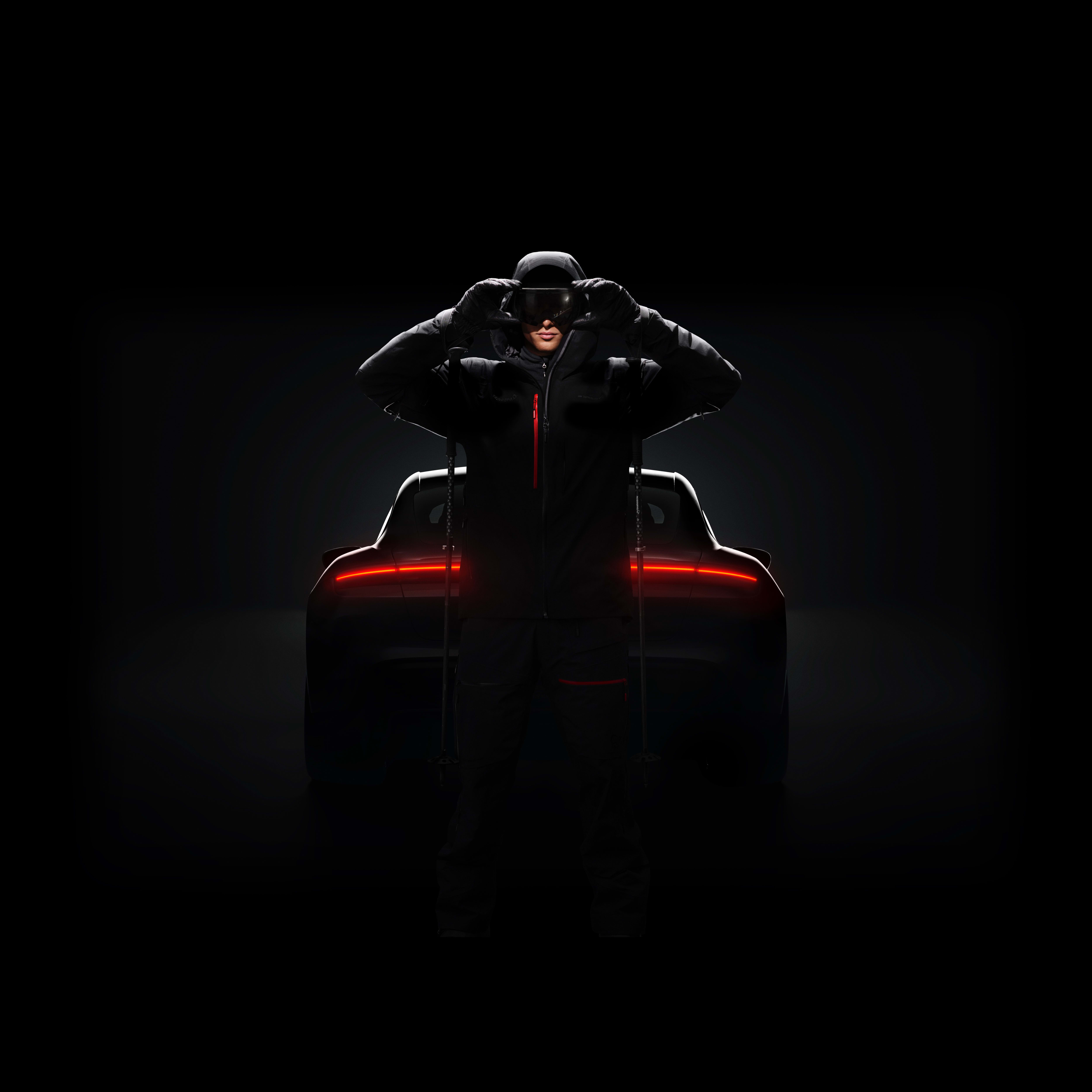 A person in black Norrøna outerwear adjusts ski goggles, ski poles visible. The taillights of a car are in the background.