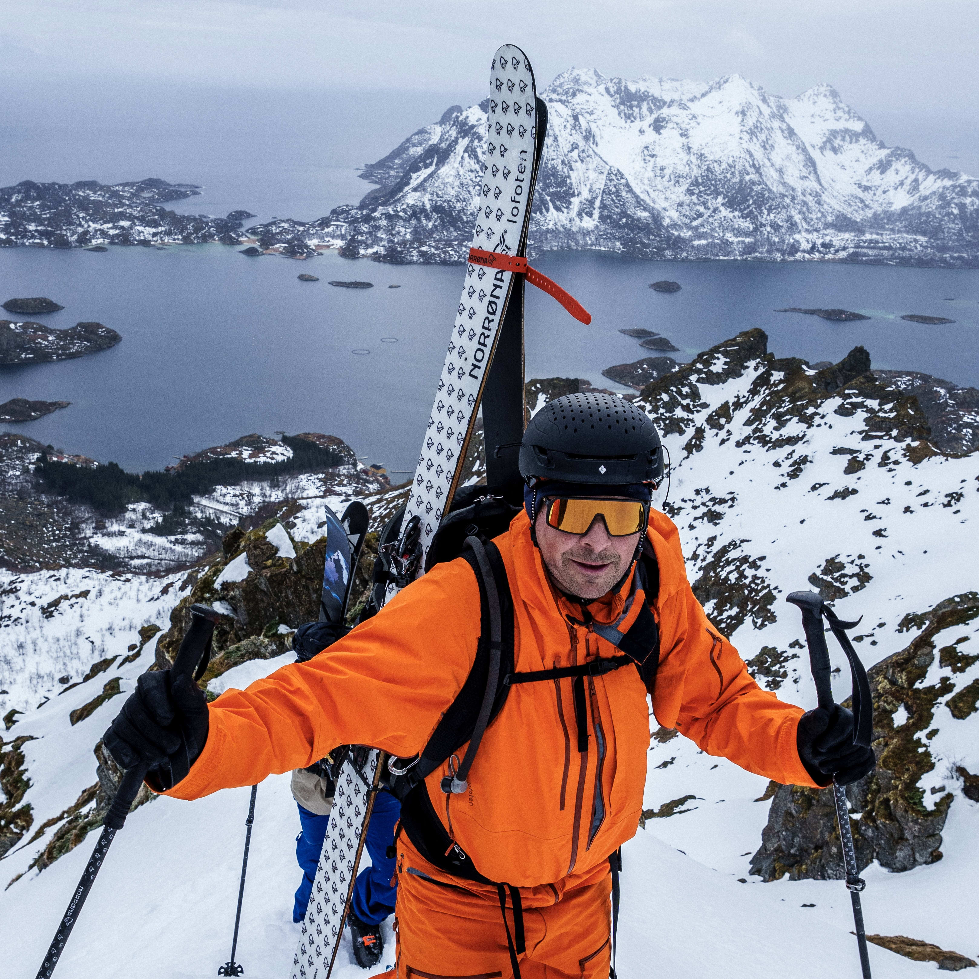 A man wearing a bright orange Norrøna ski suit, black ski gloves, a black helmet, and orange tinted goggles stands in a snowy mountain pass. The background features a snowy mountainous landscape with a body of water in the distance. The man has two black ski poles and a pair of Norrøna skis strapped to his back.