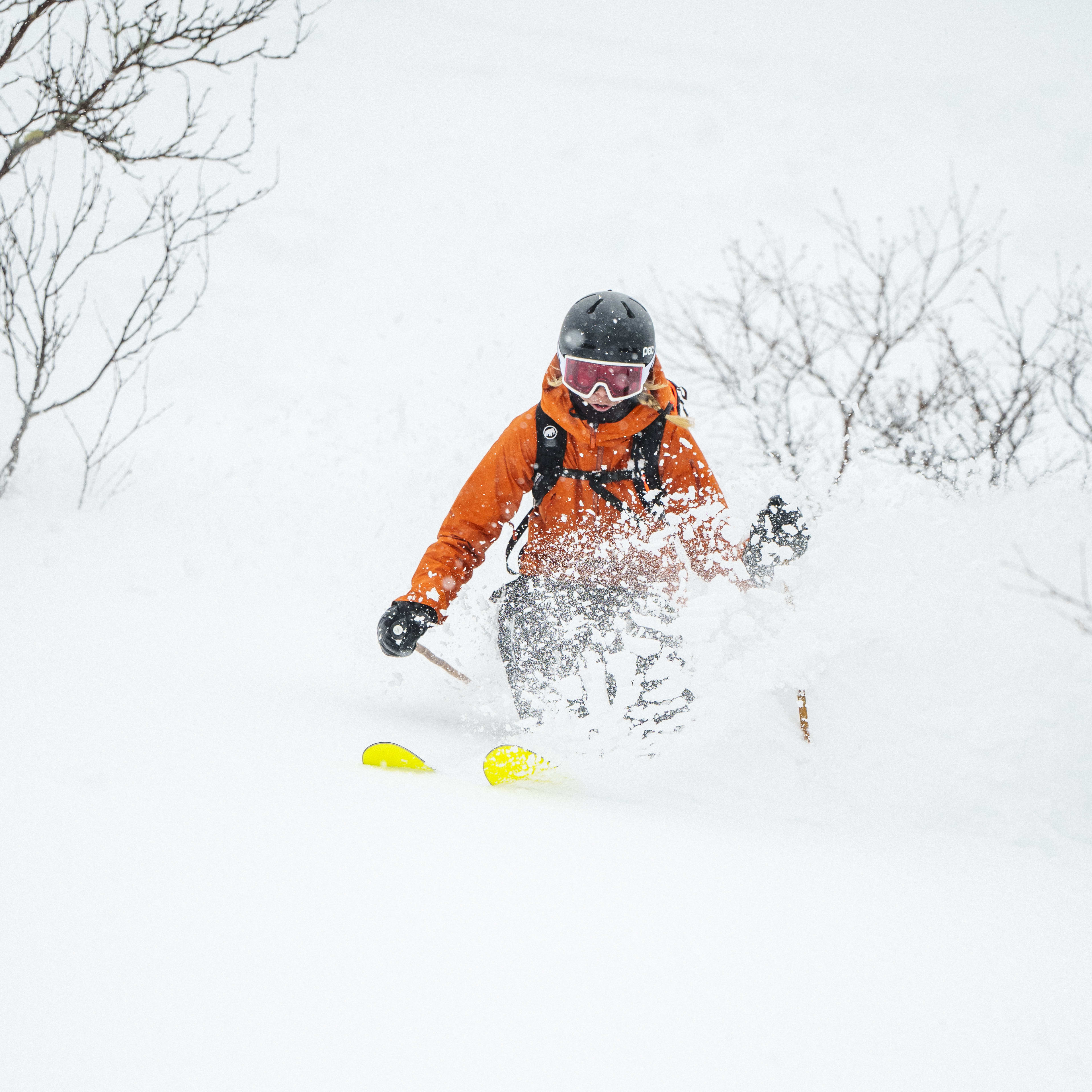 A skier in an orange Norrøna jacket is skiing down a snowy slope, leaving a trail of powder behind them.  
