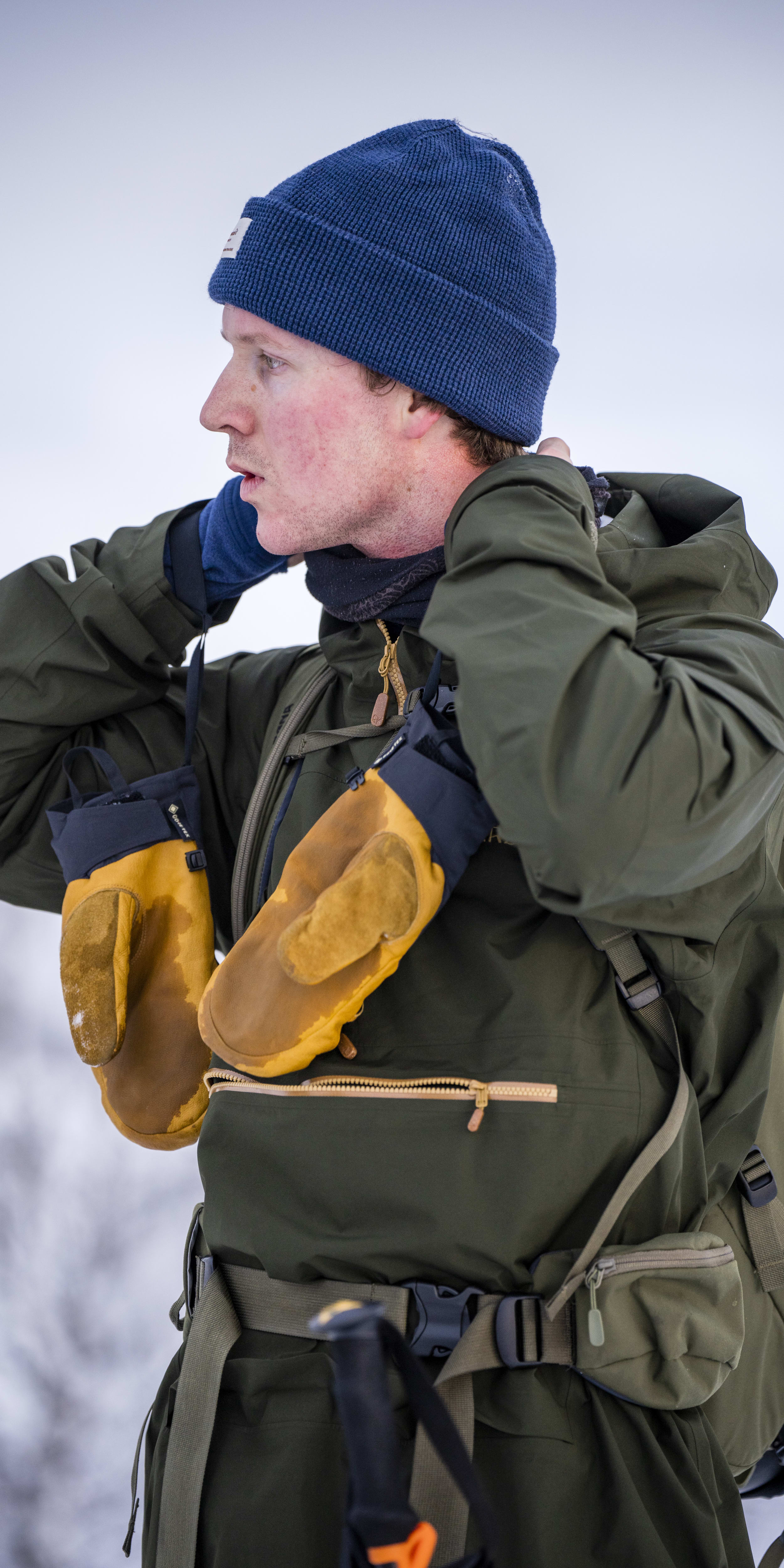 A man wearing a Norrøna jacket and gloves, a blue beanie, and a backpack. 
