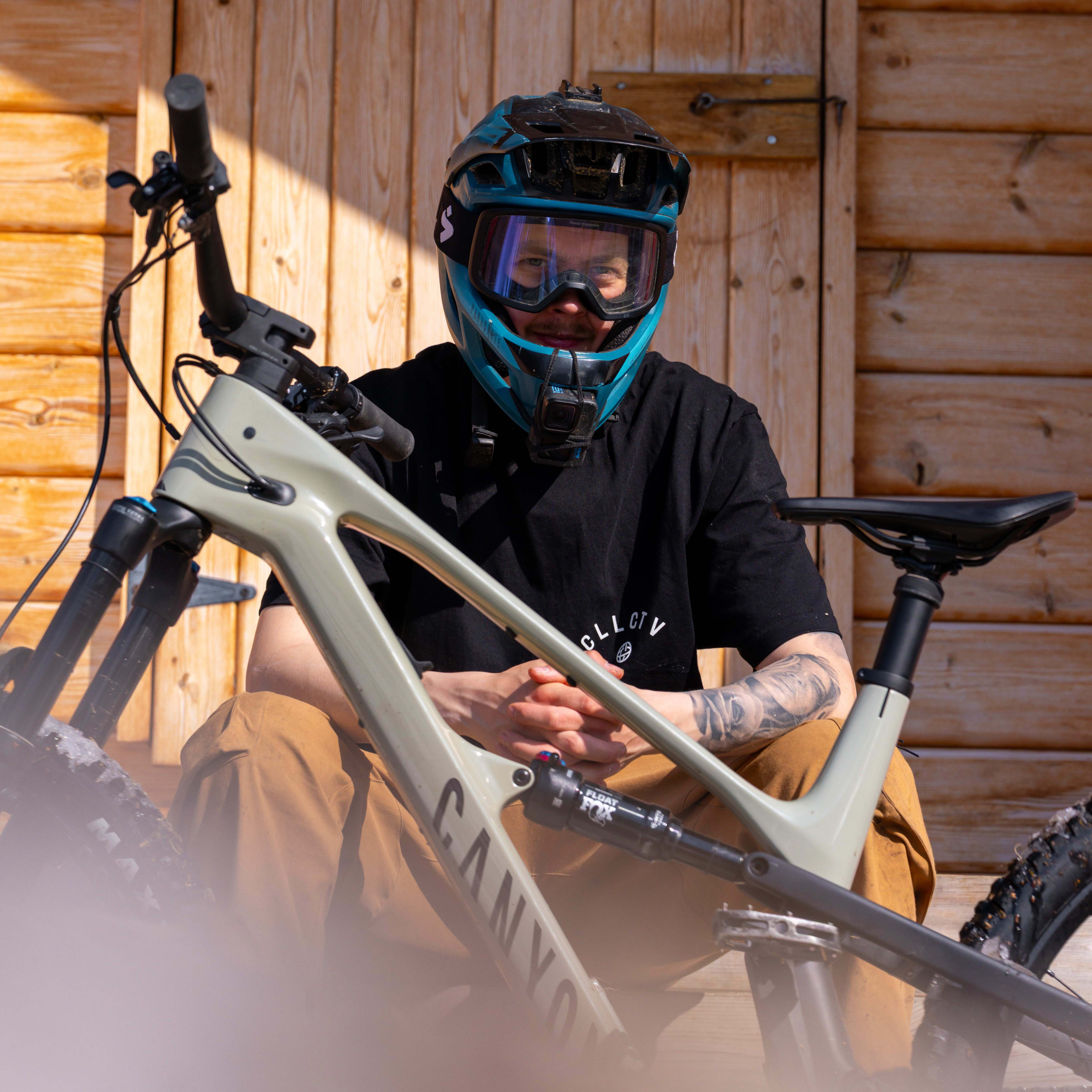 A mountain biker in full gear sits with a Canyon bike in front of a wooden building. They're wearing a Norrøna t-shirt and have a turquoise helmet and goggles.