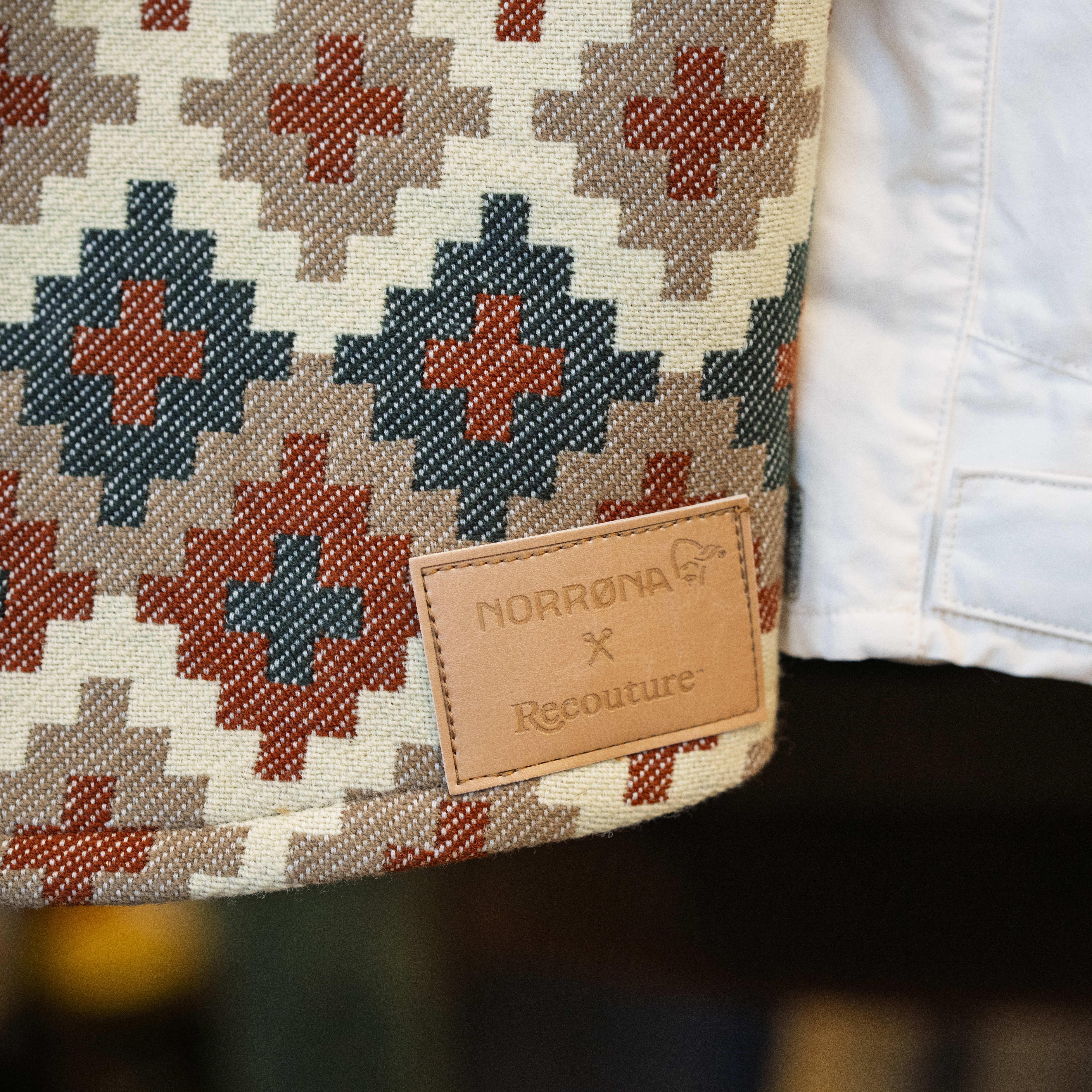 Close-up of a Norrøna garment with a geometric pattern, featuring a leather Norrøna x Recouture tag, and adjacent white clothing.
