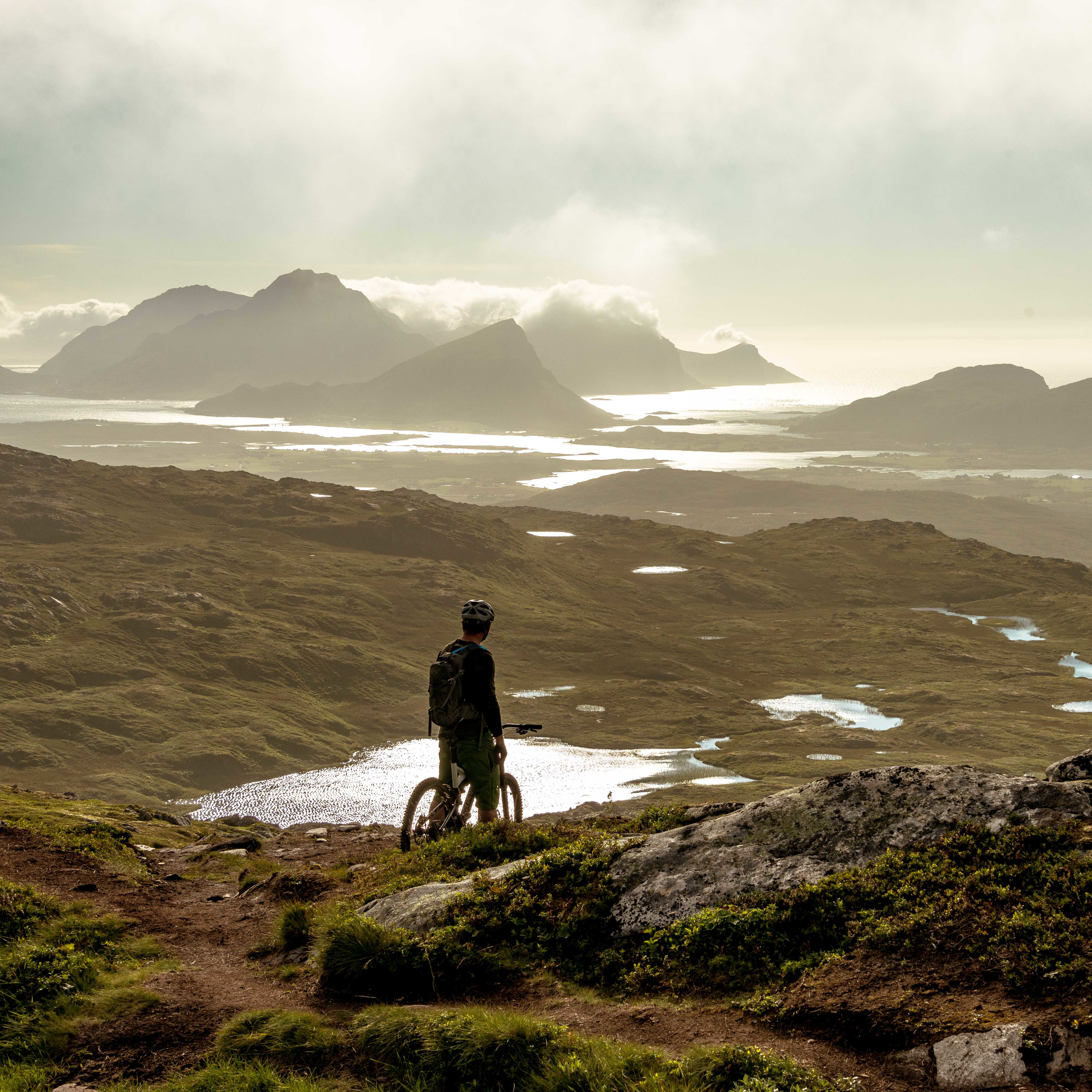 A person standing on a mountain top overlooking a valley and ocean. The person is wearing a Norrøna backpack and is holding a bike.