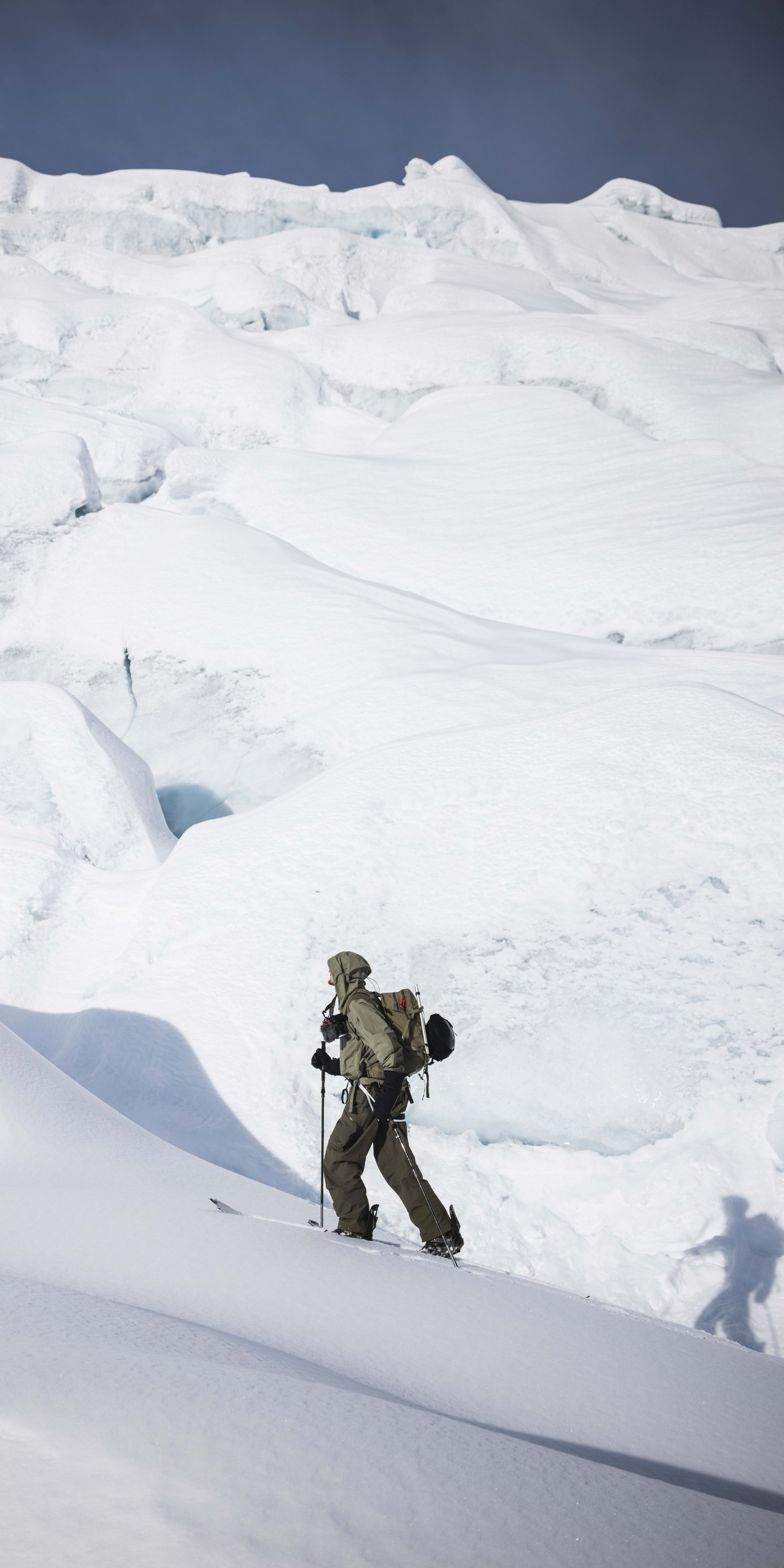 A hiker with a backpack and Norrøna clothing uses ski poles to ascend a snow-covered mountain with a large glacier in the background.