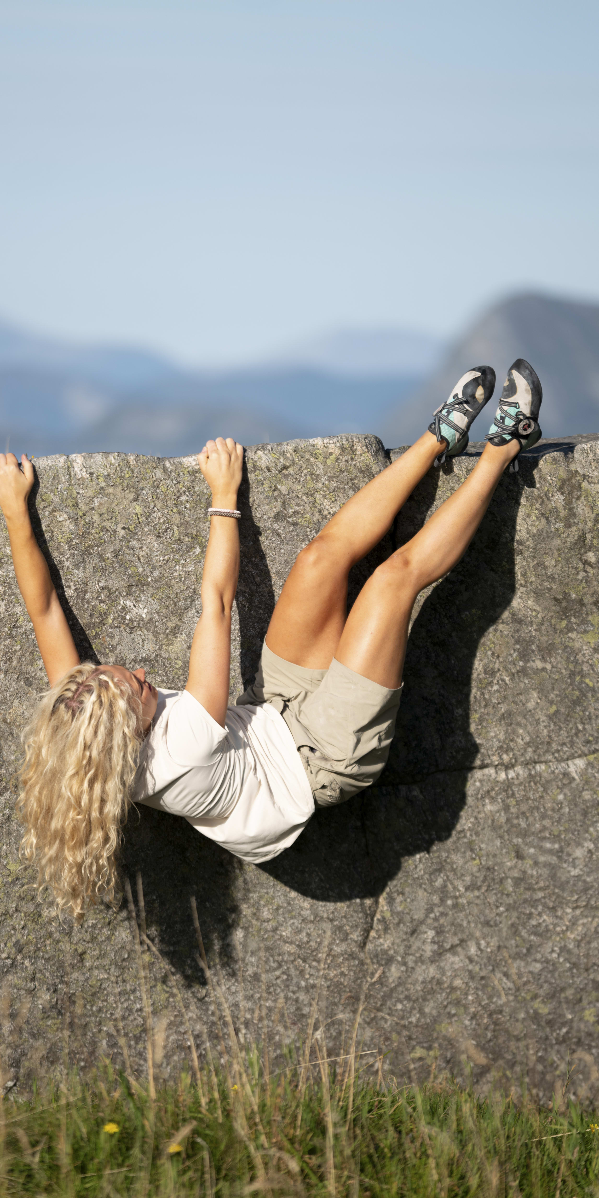 A woman with blonde hair hangs from a rock, wearing a white t-shirt and Norrøna shorts. Mountains are visible in the background.