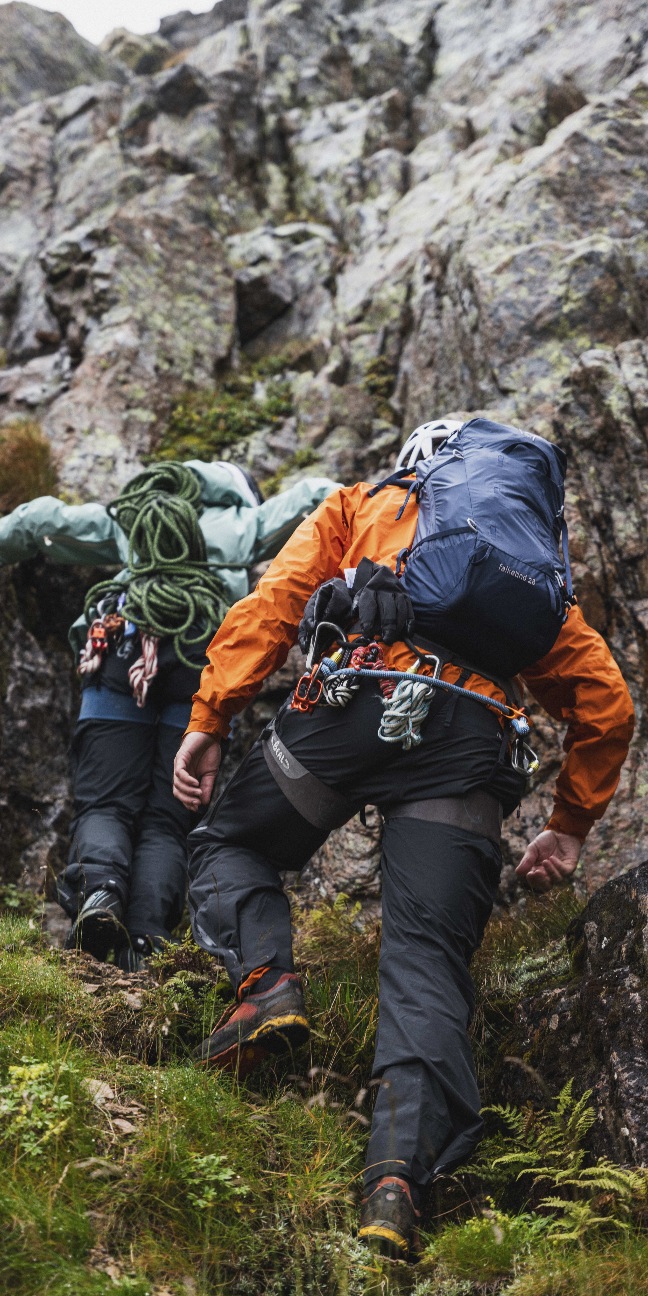 Two people wearing Norrøna gear climb a rocky mountainside. They are wearing helmets, climbing harnesses, and carrying rope.  
