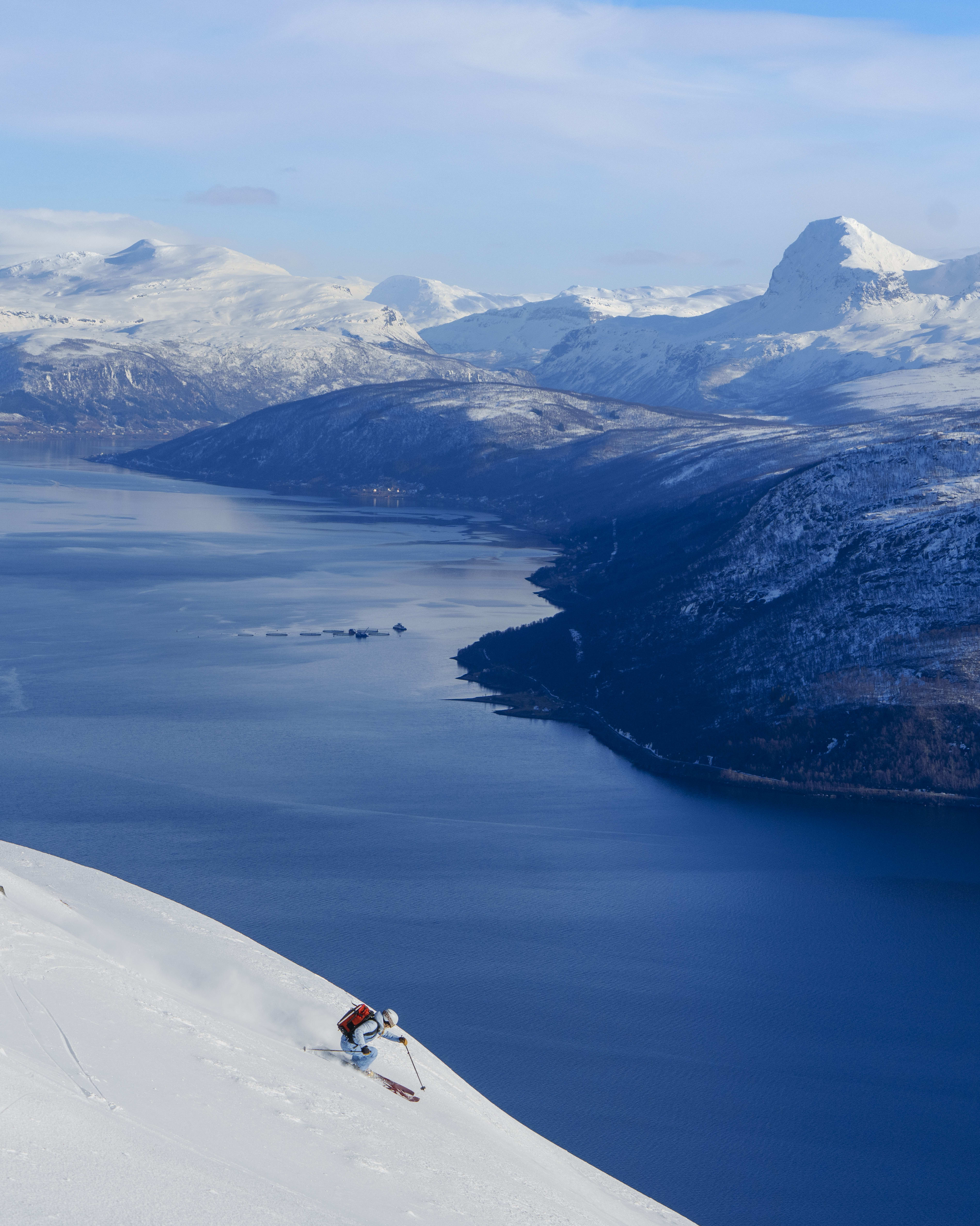 A skier wearing Norrøna clothing skis down a snowy slope towards a blue fjord. Snow-capped mountains rise in the background under a bright sky.