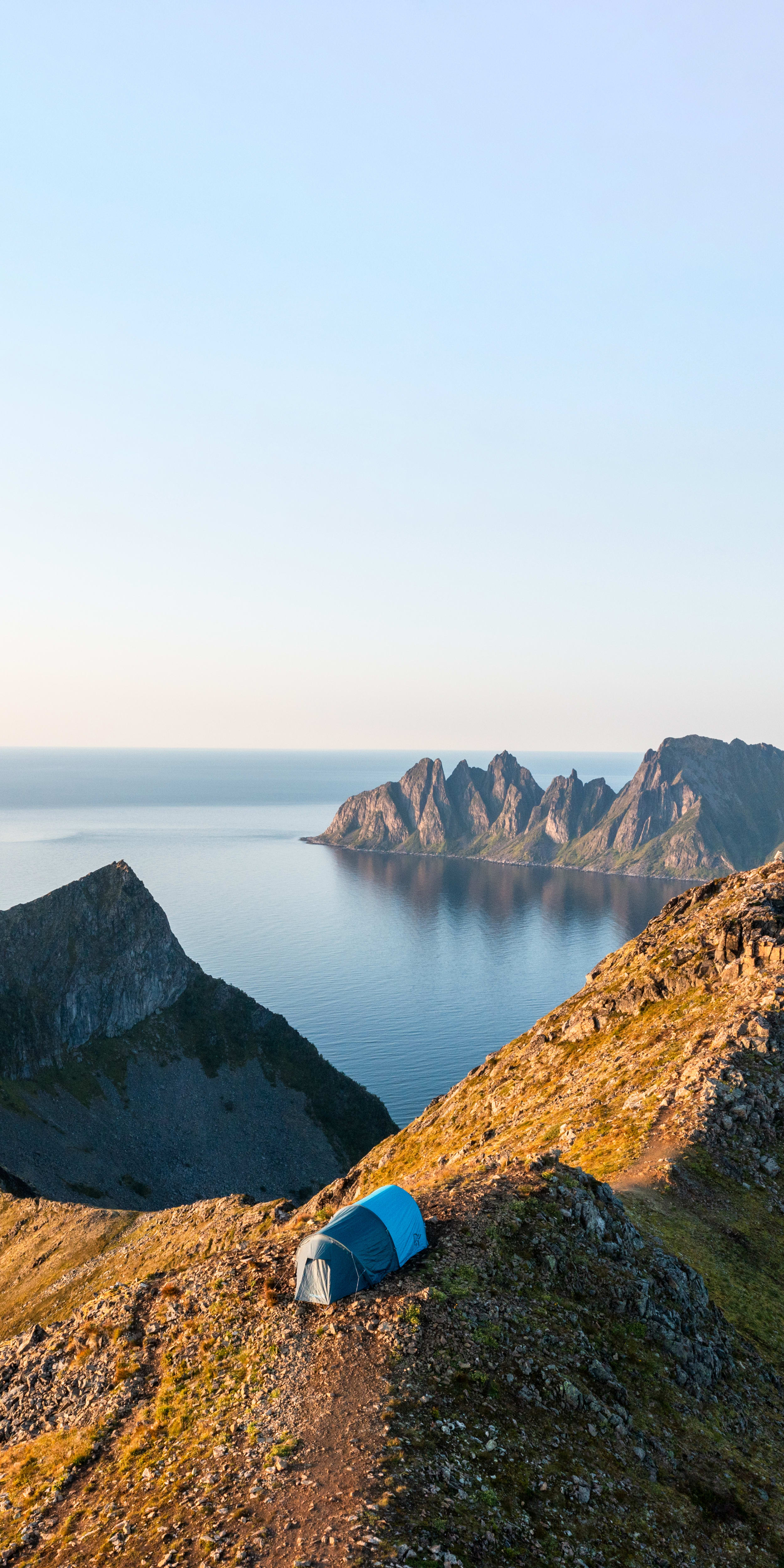 A blue Norrøna tent sits on a mountain peak overlooking a fjord. 

