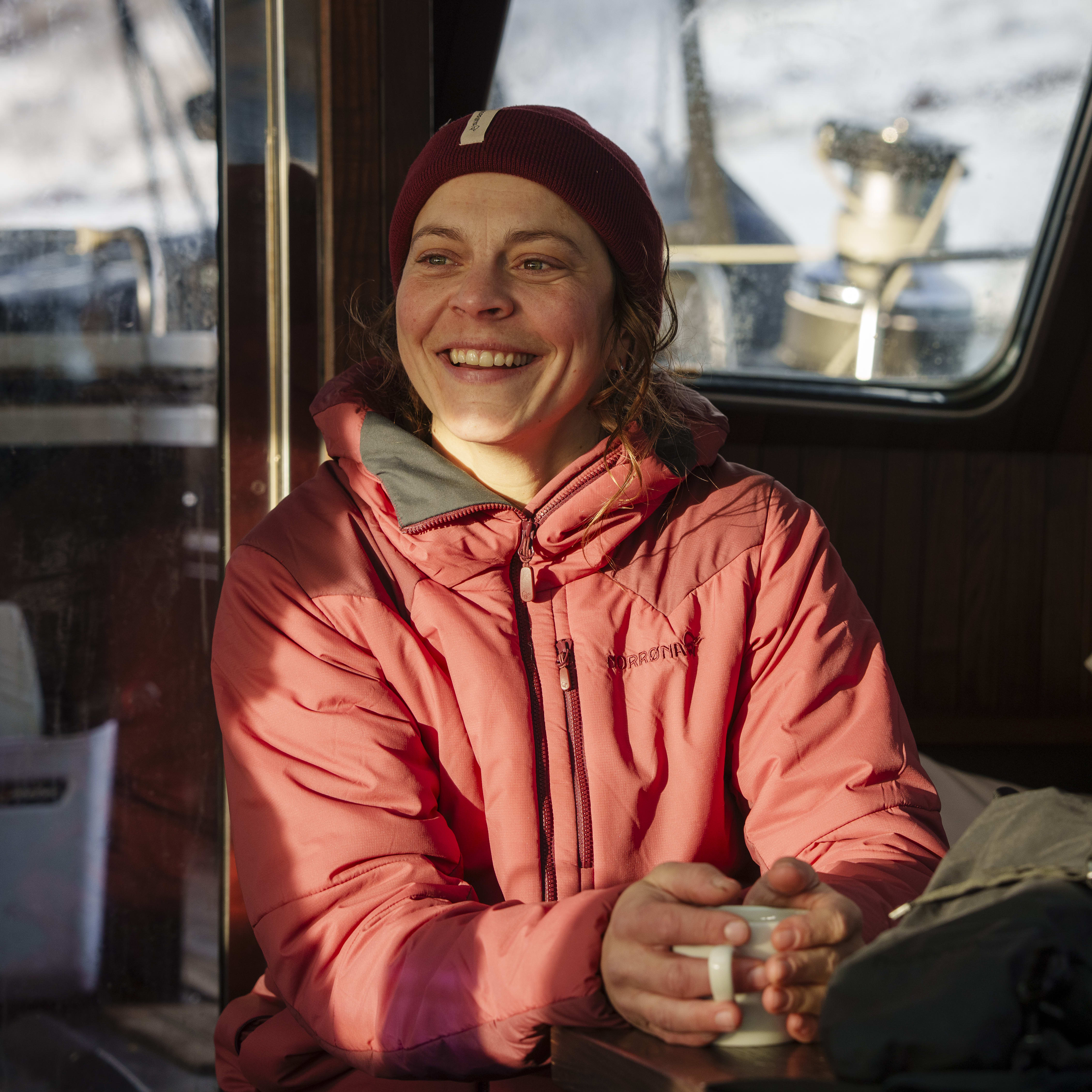 A smiling woman wearing a Norrøna jacket and hat sits on a boat, looking out the window.