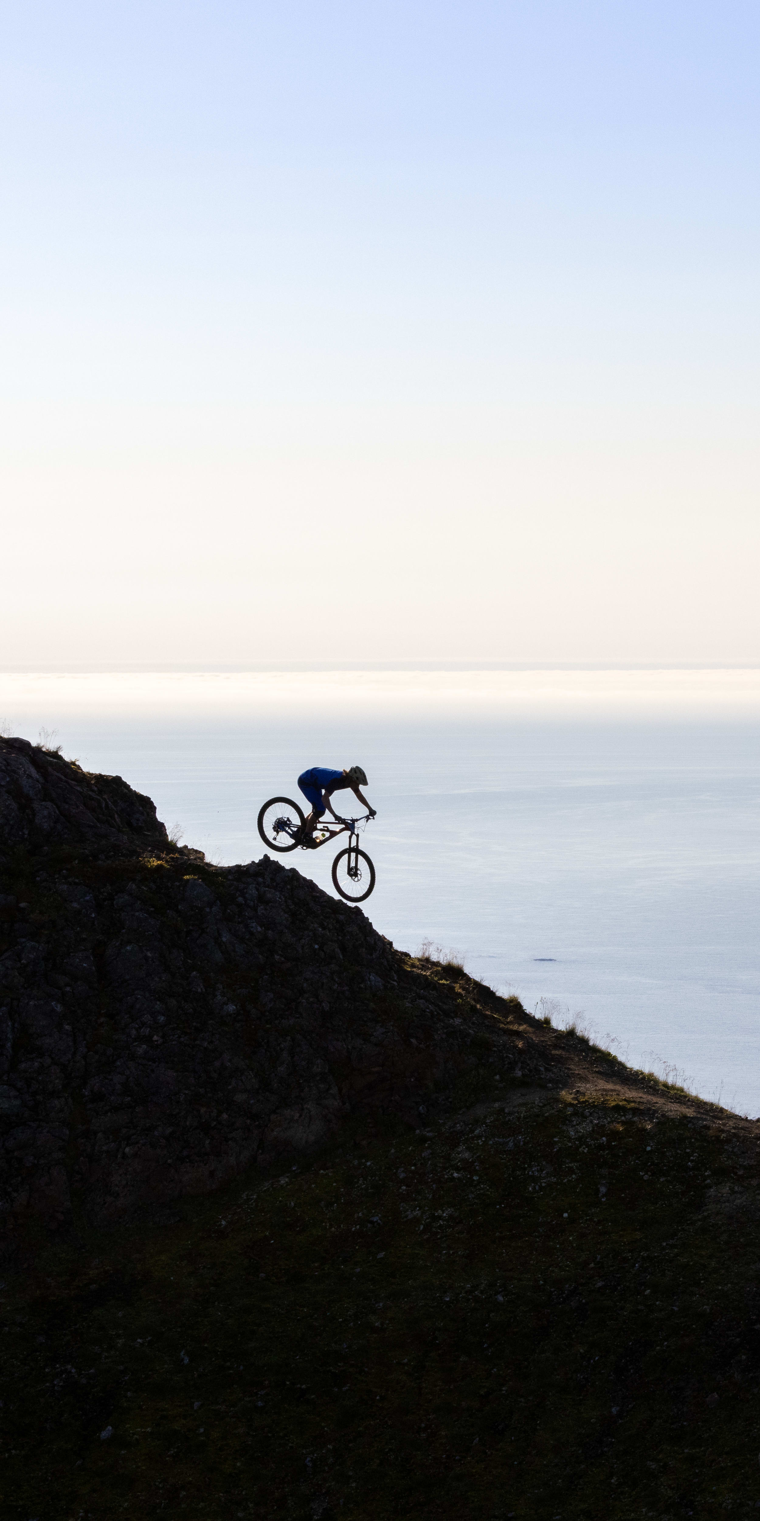 Silhouette of a person riding a Norrøna mountain bike down a rocky cliff overlooking the ocean. 
