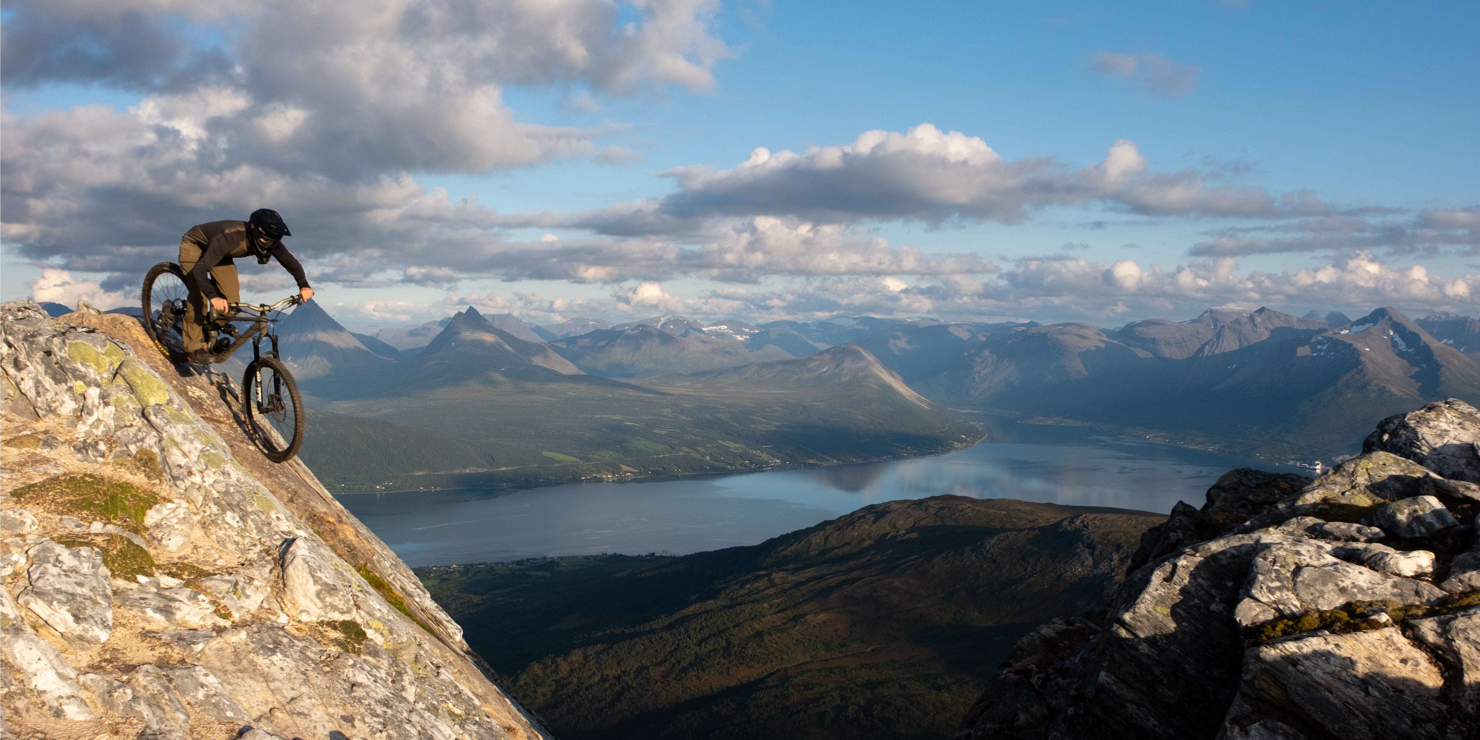 A mountain biker in Norrøna clothing navigates a rocky peak, set against a stunning mountain and fjord landscape under a cloudy sky.
