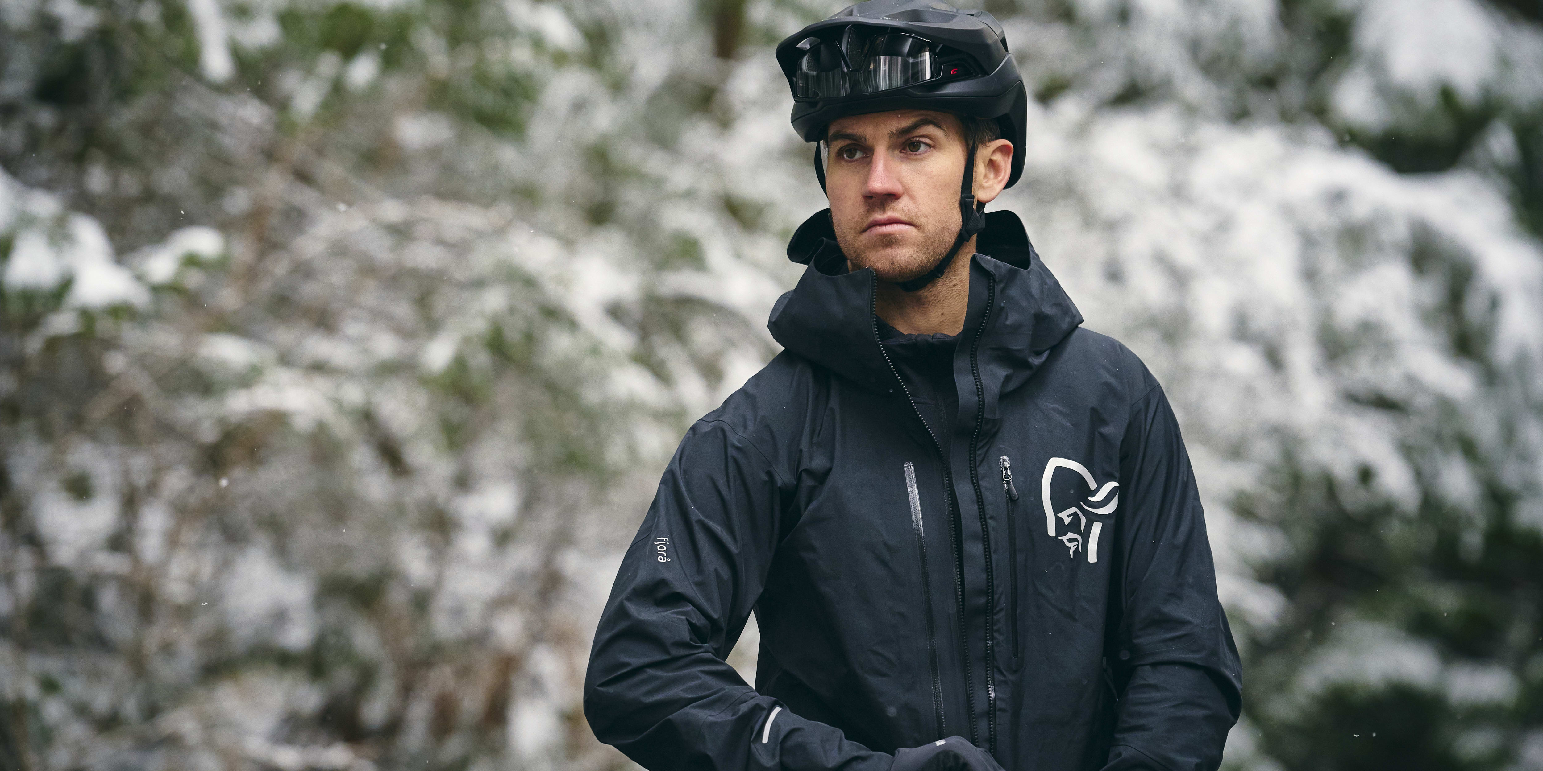 A man wearing a black Norrøna jacket and a black helmet in a snowy forest.