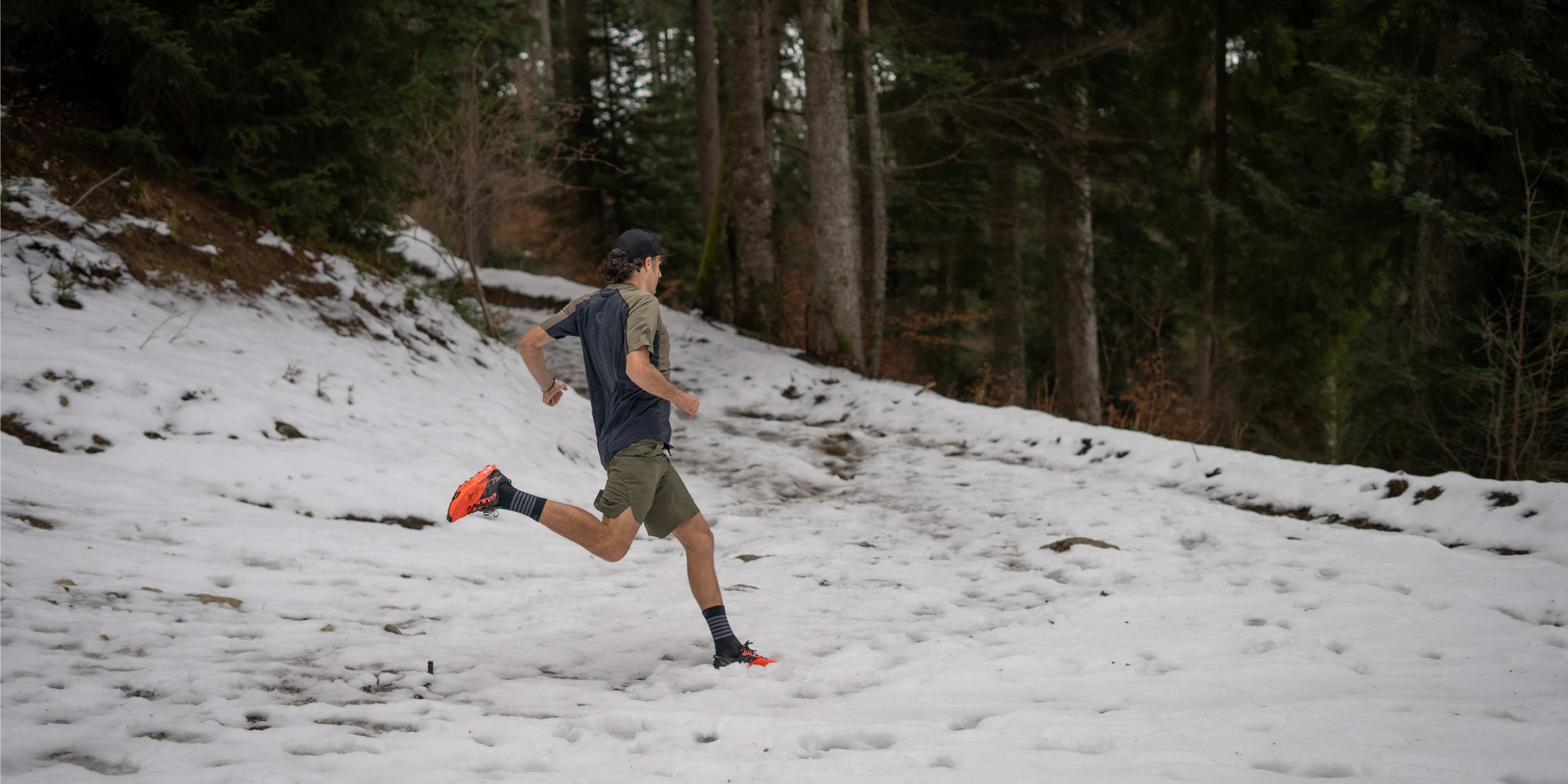 A man wearing a Norrøna shirt and shorts runs through a snowy forest. 
