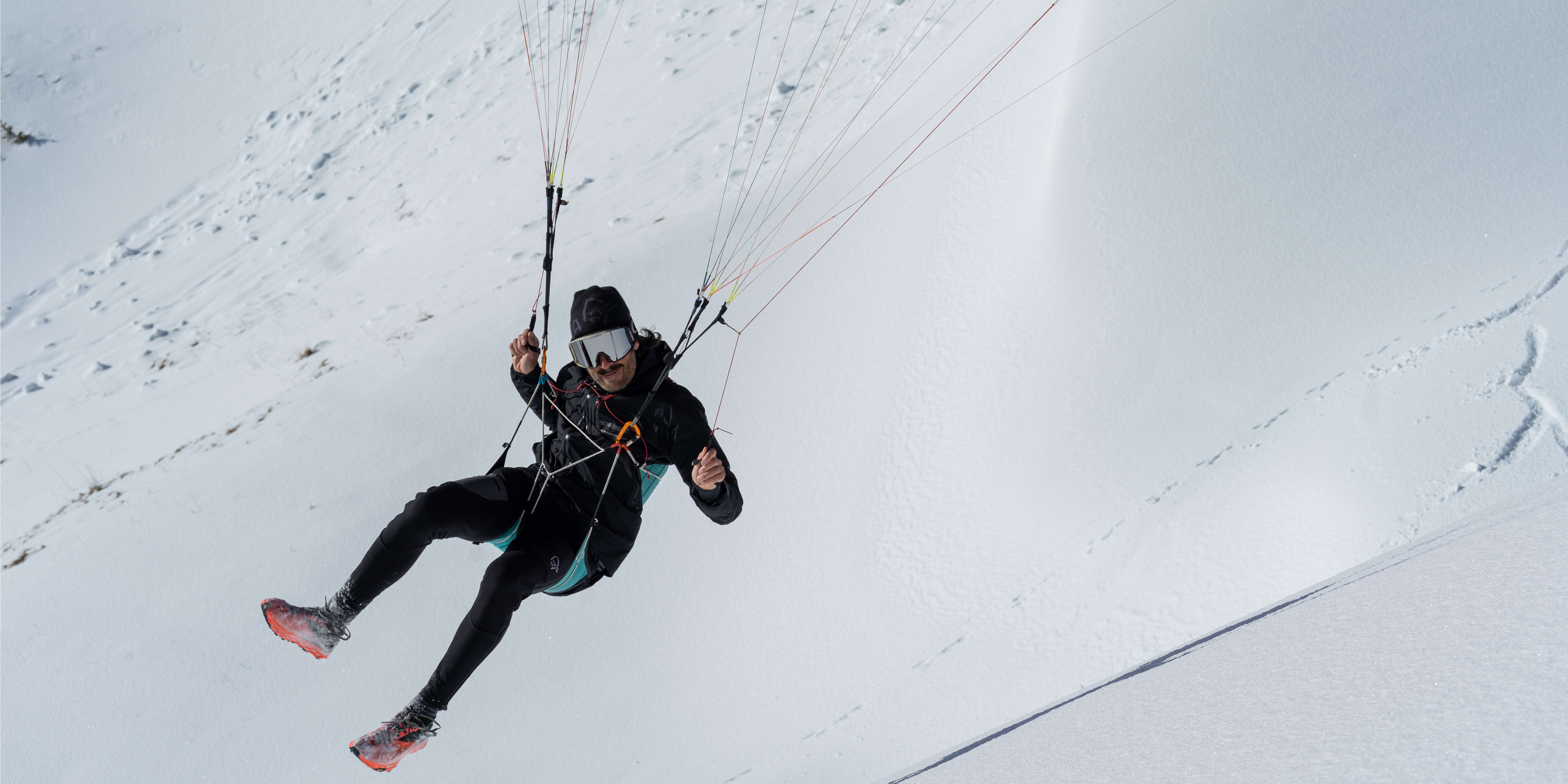 A person wearing a black jacket, a helmet, and goggles is paragliding over a snowy landscape.  The person is smiling.  The paraglider is attached to the person with ropes.  The snowy landscape is in the background.