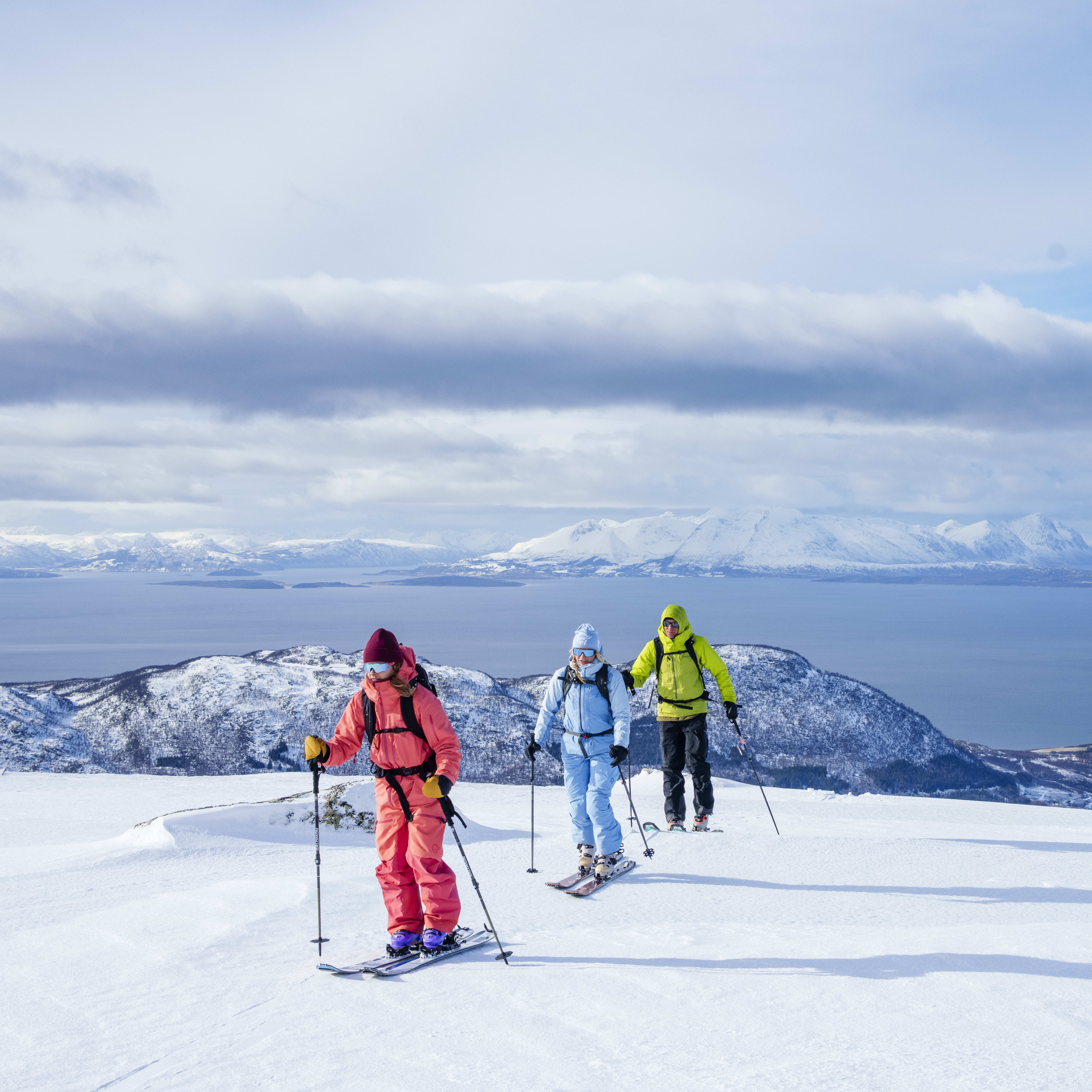 Three skiers wearing Norrøna gear climb a snowy mountain. A blue sky, islands, and ocean are in the background.
