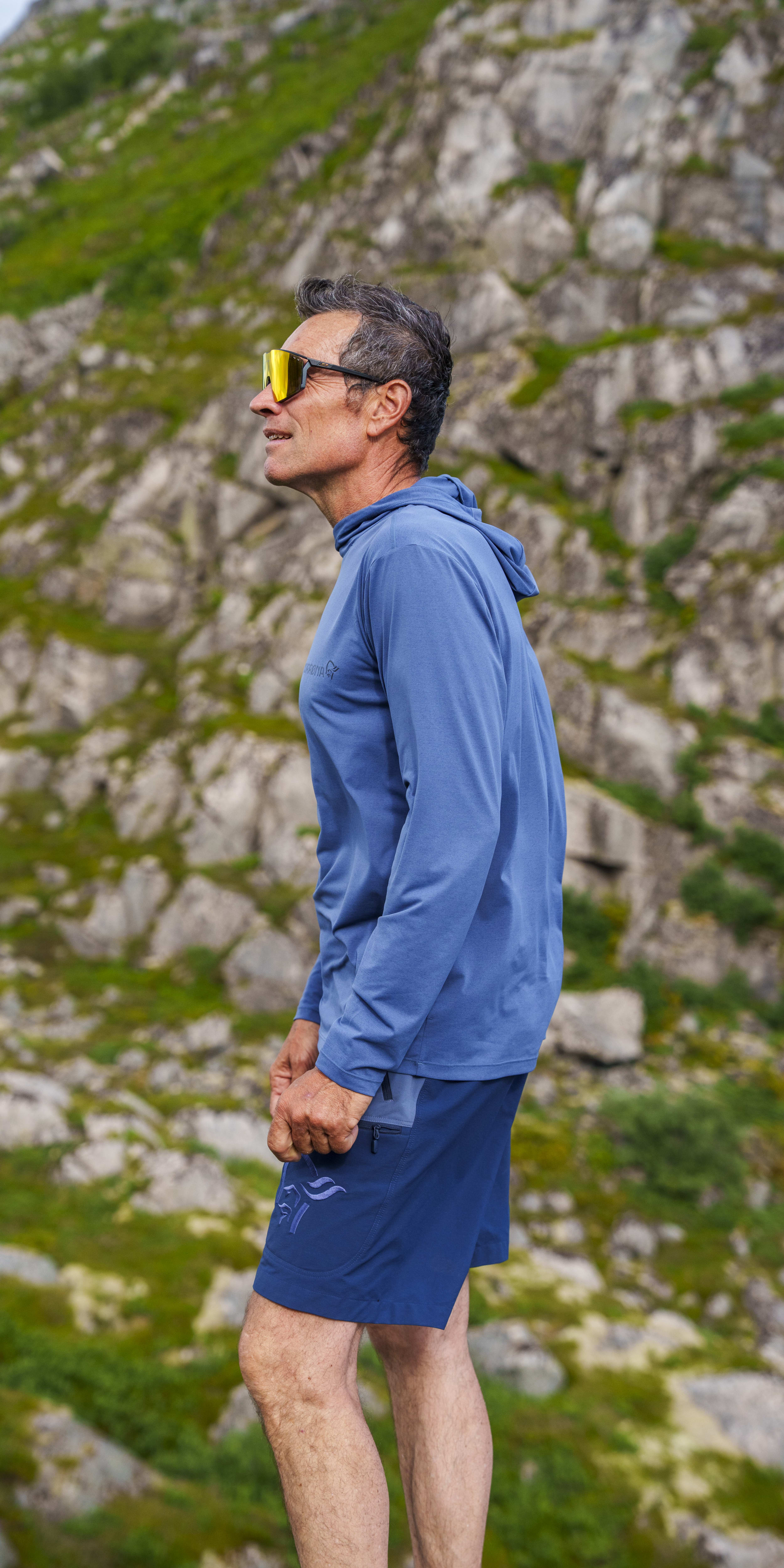 Man in Norrøna hoodie and shorts, yellow sunglasses, standing sideways against a mountain background.