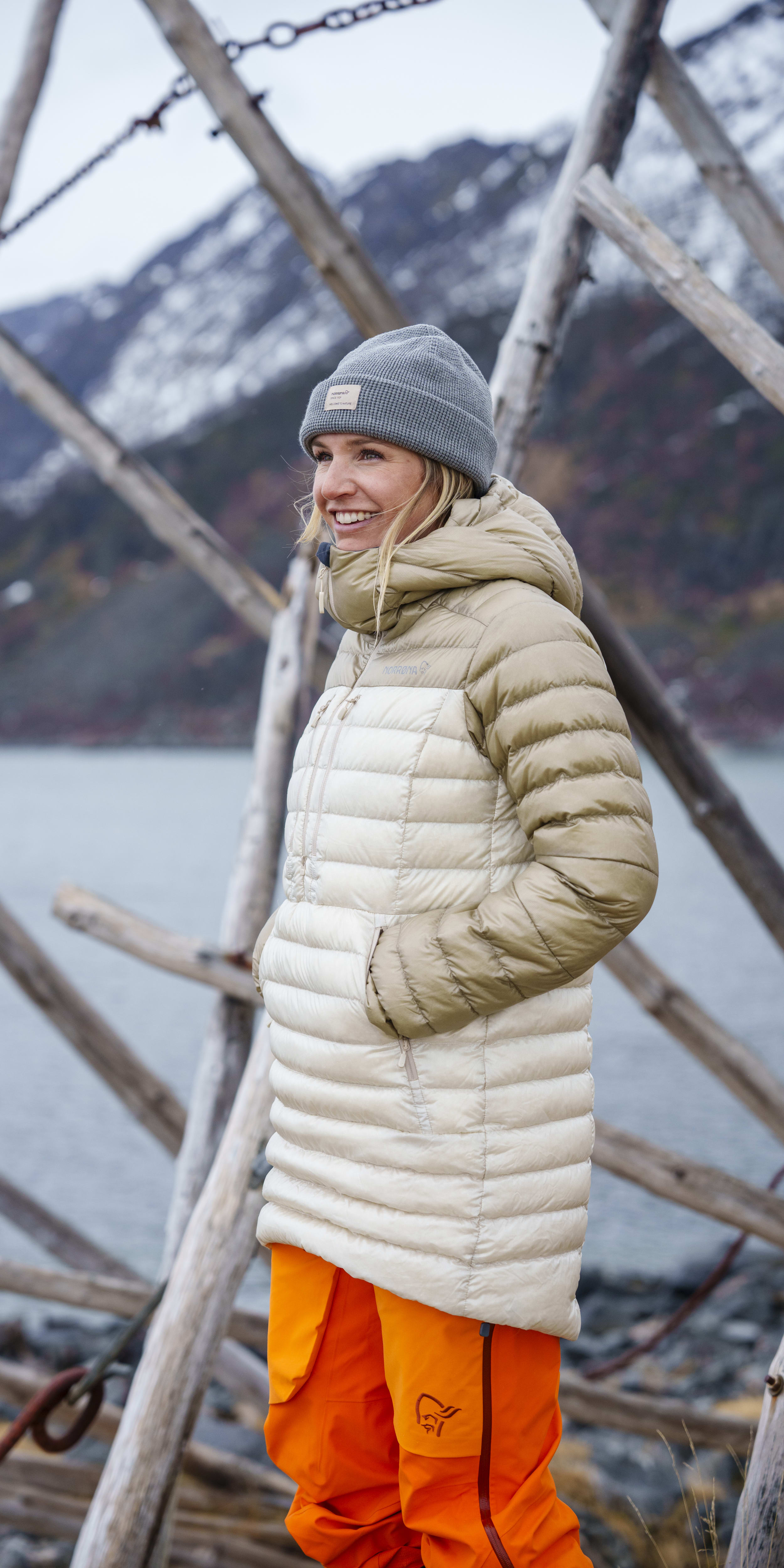 A woman wearing a gray beanie, a white down jacket, and orange pants is smiling and standing in front of a wooden structure near a body of water.