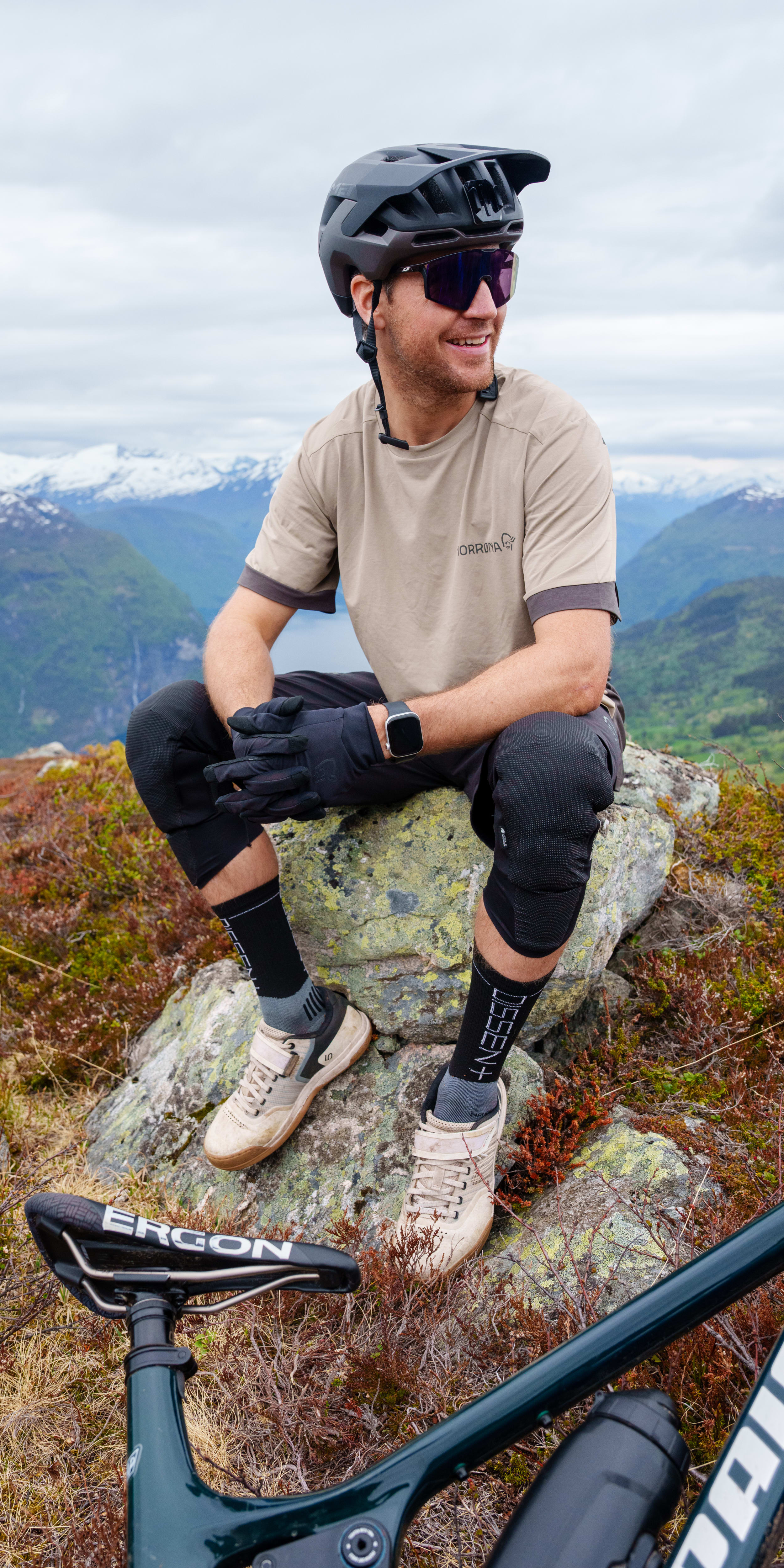 Mountain biker in Norrøna gear sitting on a rock in a scenic mountain landscape.