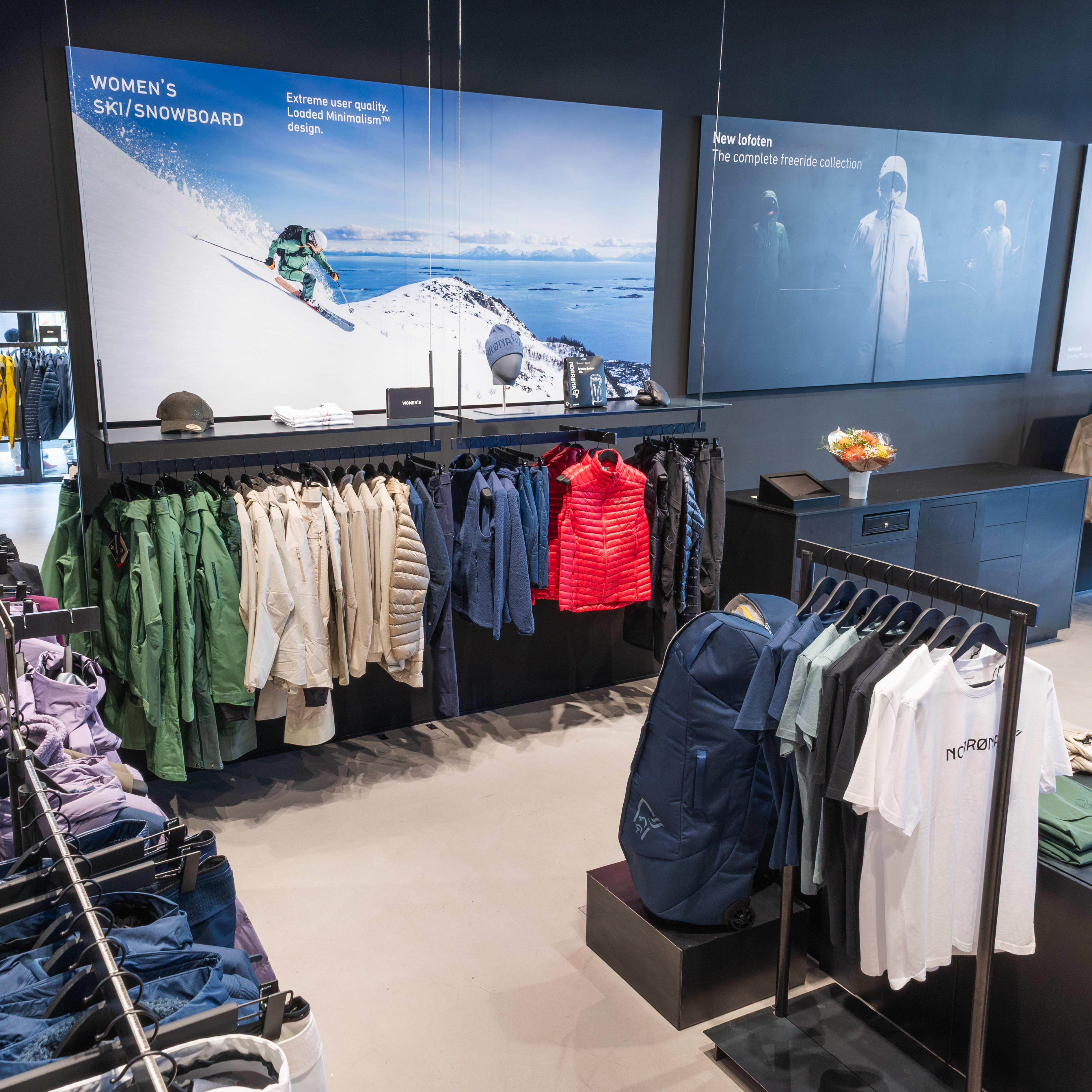 Store interior with Norrøna clothing on racks and displays. Includes jackets, pants, shirts, and a backpack. Winter sports posters on the back wall.