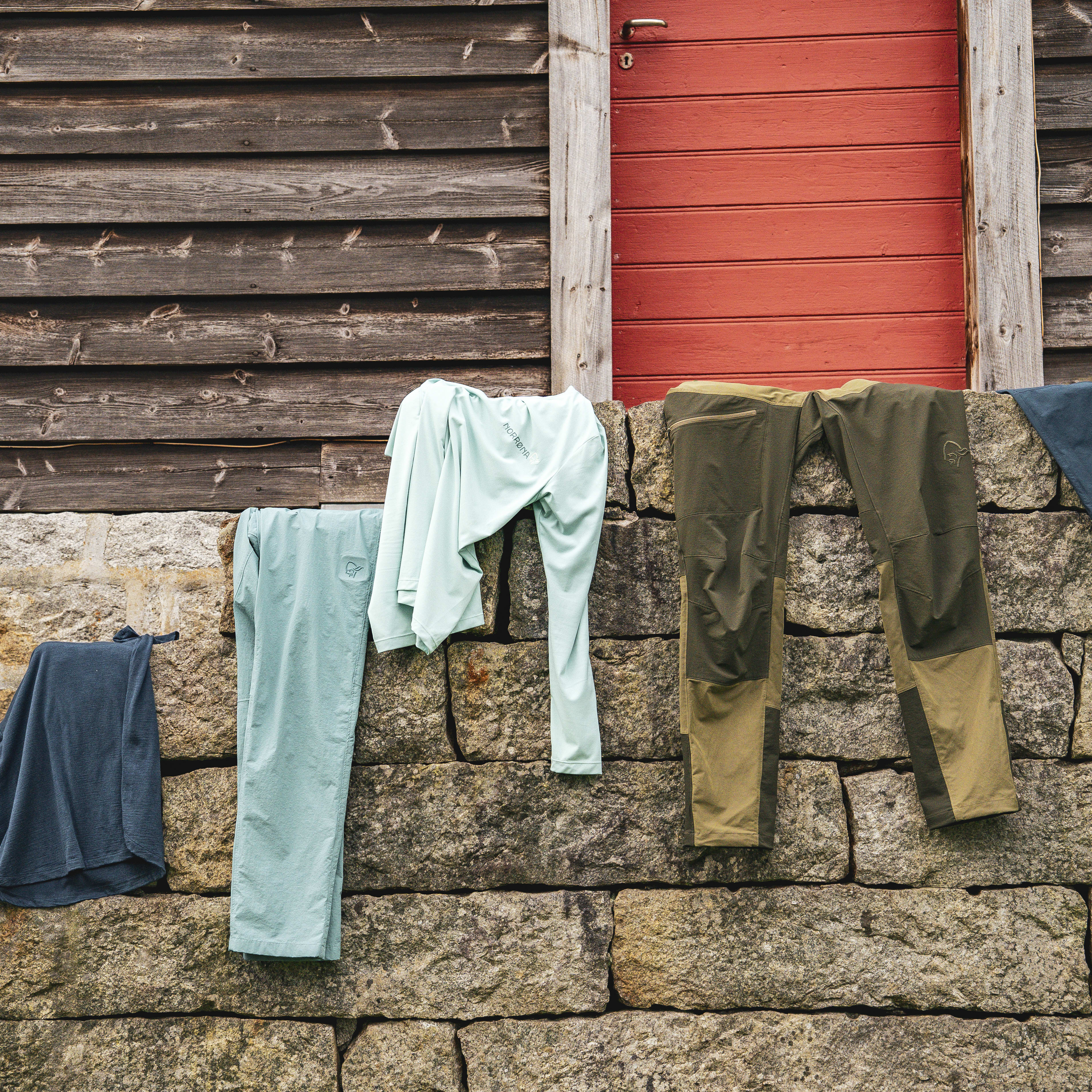 Norrøna apparel hangs on a stone wall outside a wooden building with a red door and window.