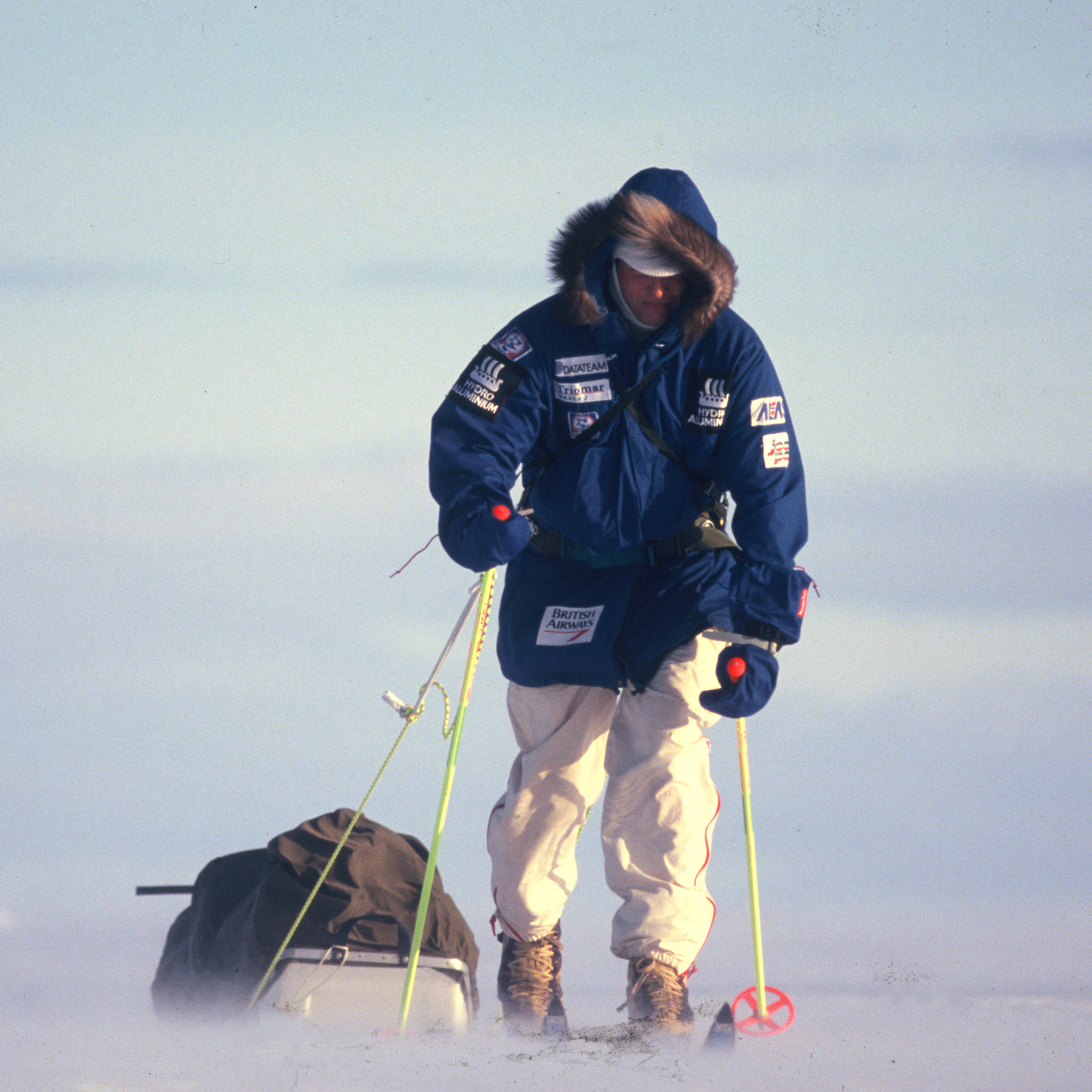 A person in a blue winter jacket and white pants is pulling a sled across snowy terrain. They are wearing a white hat and gloves and using two ski poles to propel themselves forward.