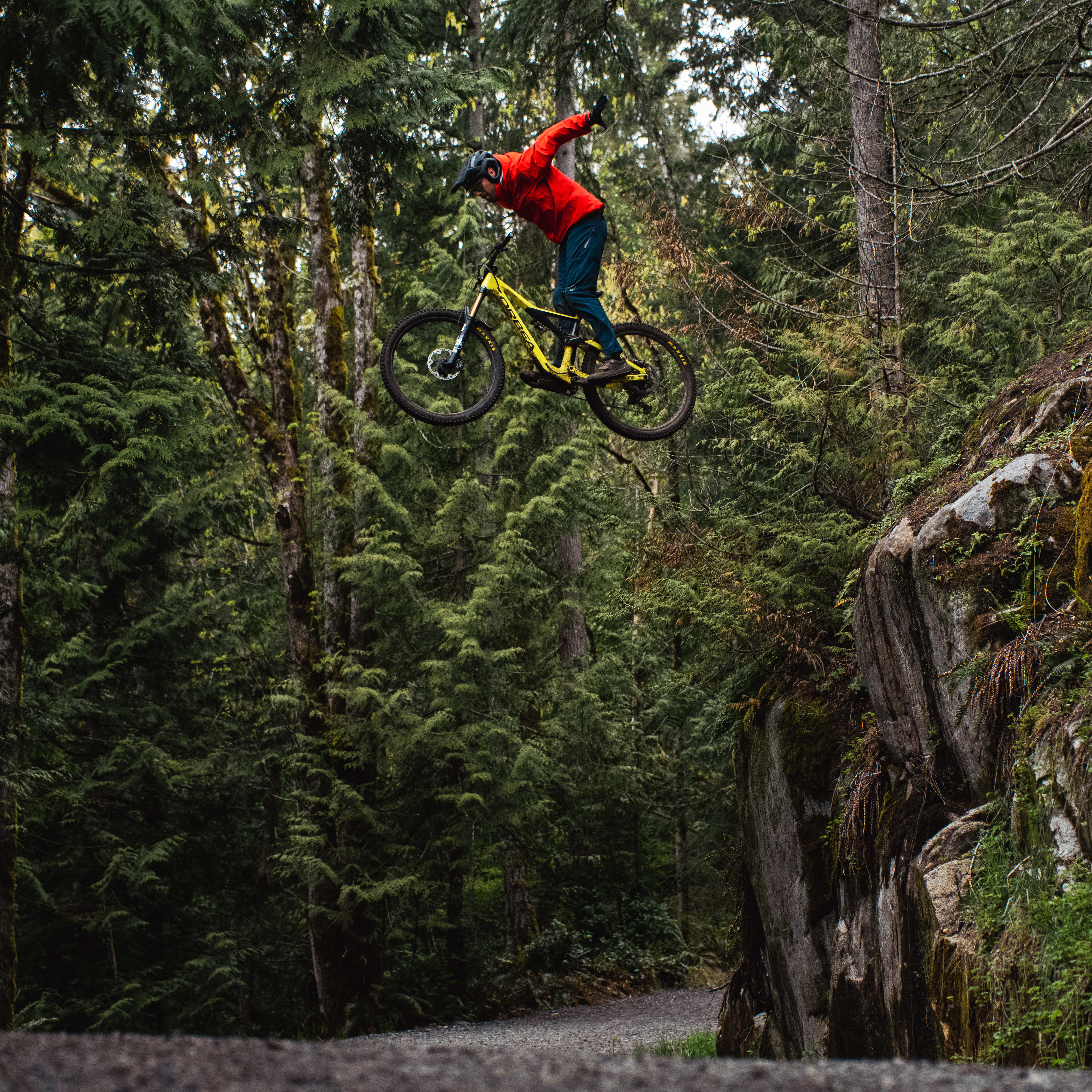 A person wearing a Norrøna jacket jumps high on a yellow mountain bike in a green forest.