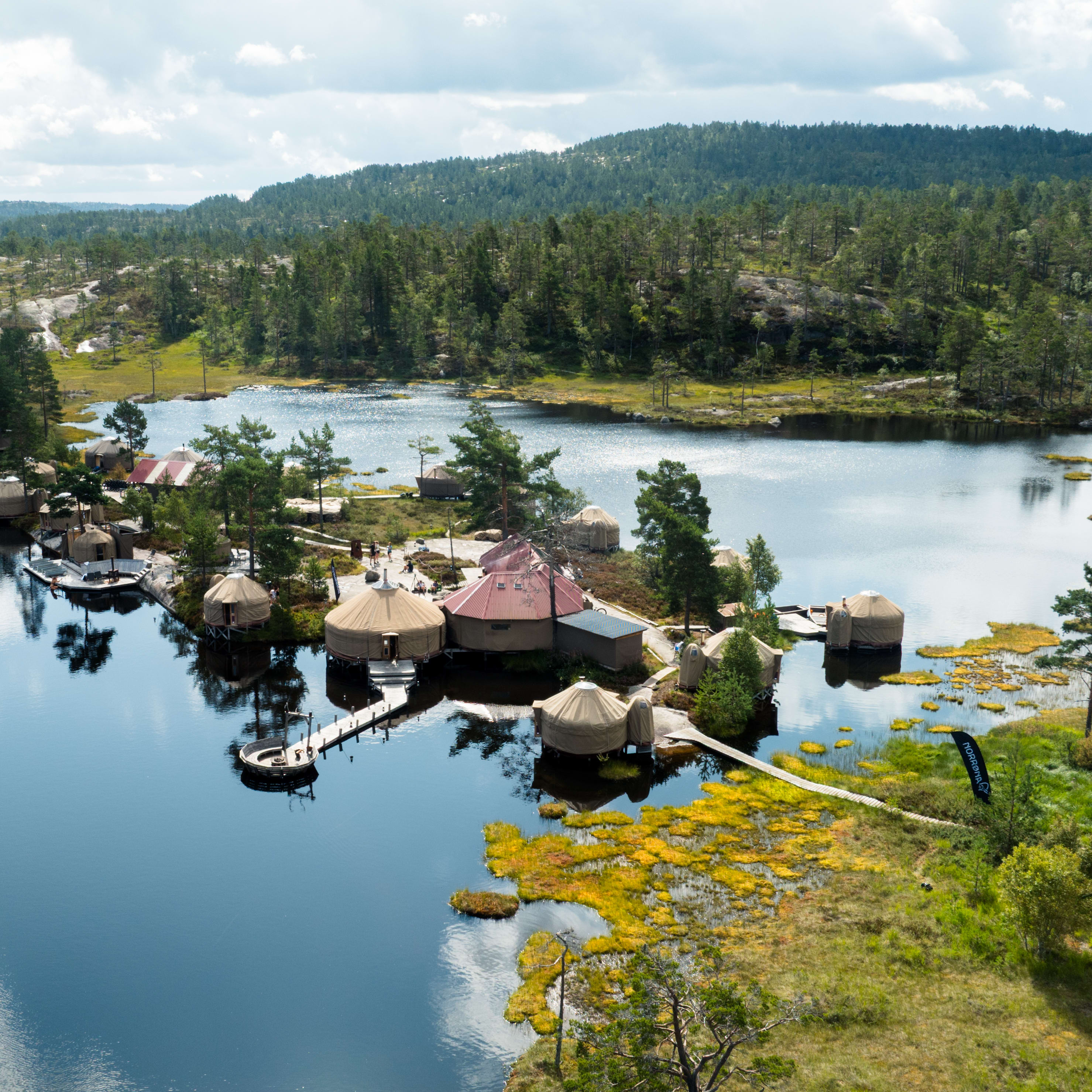 Aerial view of a lakeside resort with yurt-style accommodations, surrounded by forest and wetlands under a cloudy sky. Norrøna flag is visible.
