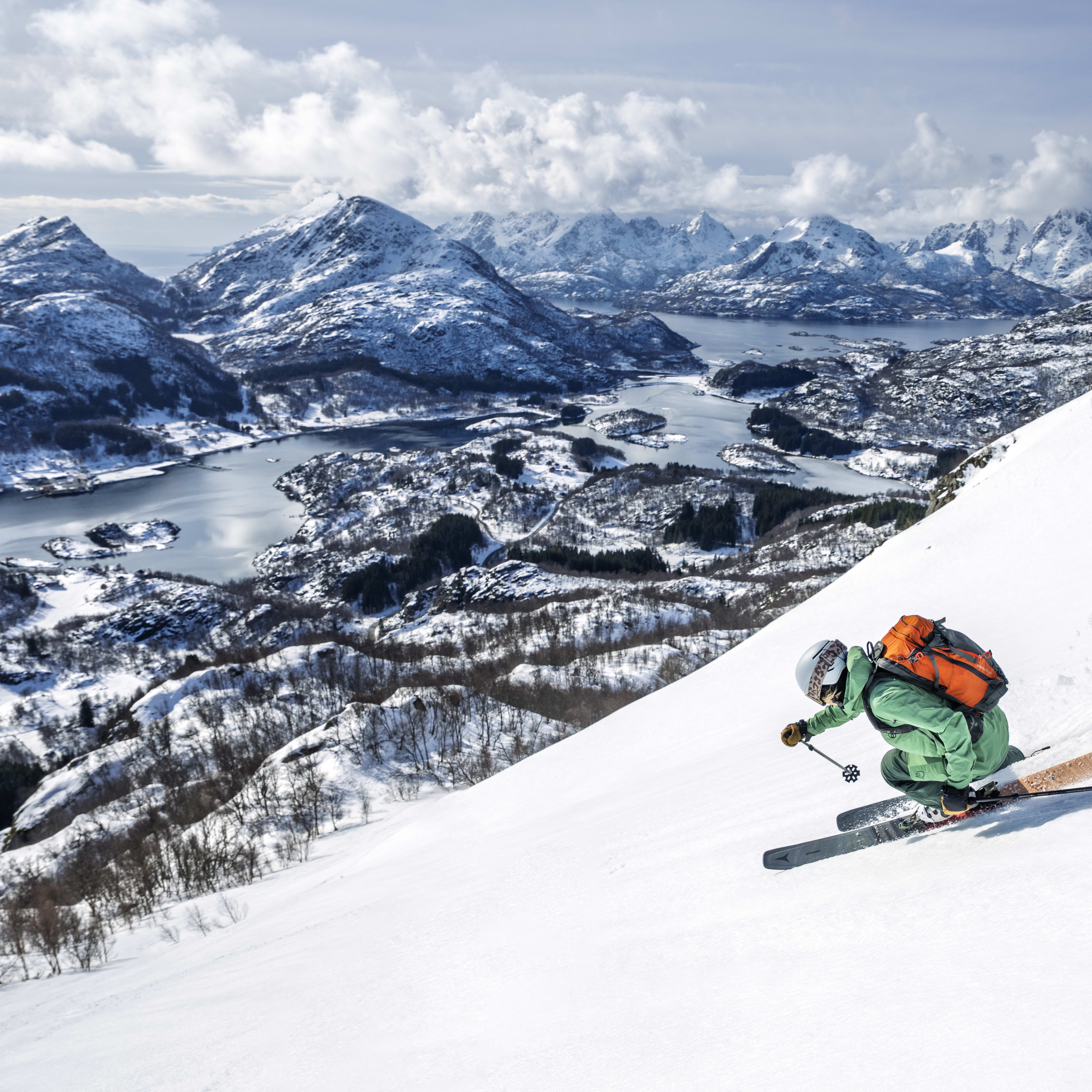 A skier wearing Norrøna clothing descends a snowy mountain slope with a view of a fjord and mountains in the background.