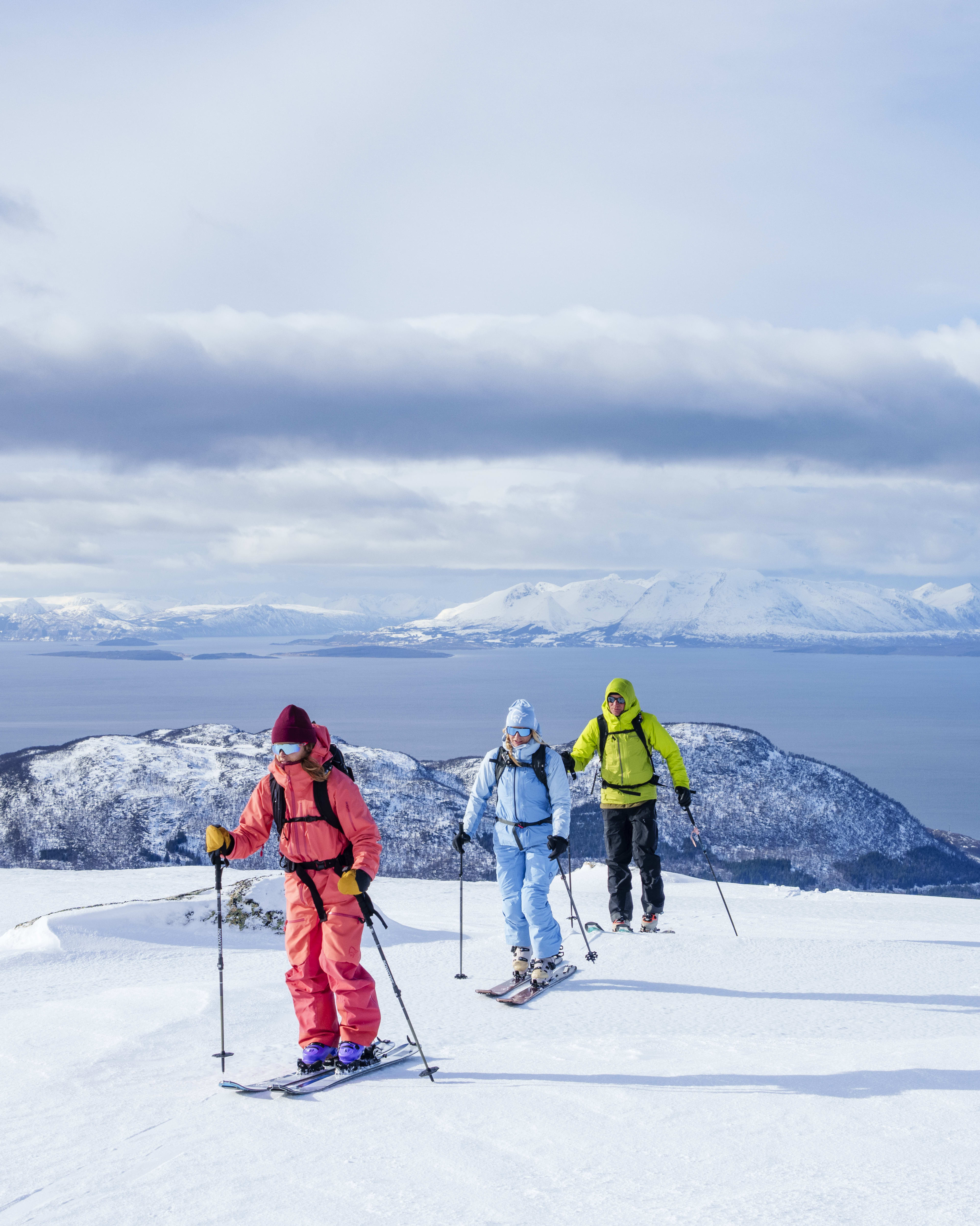 Three skiers wearing Norrøna gear climb a snowy mountain. A blue sky, islands, and ocean are in the background.