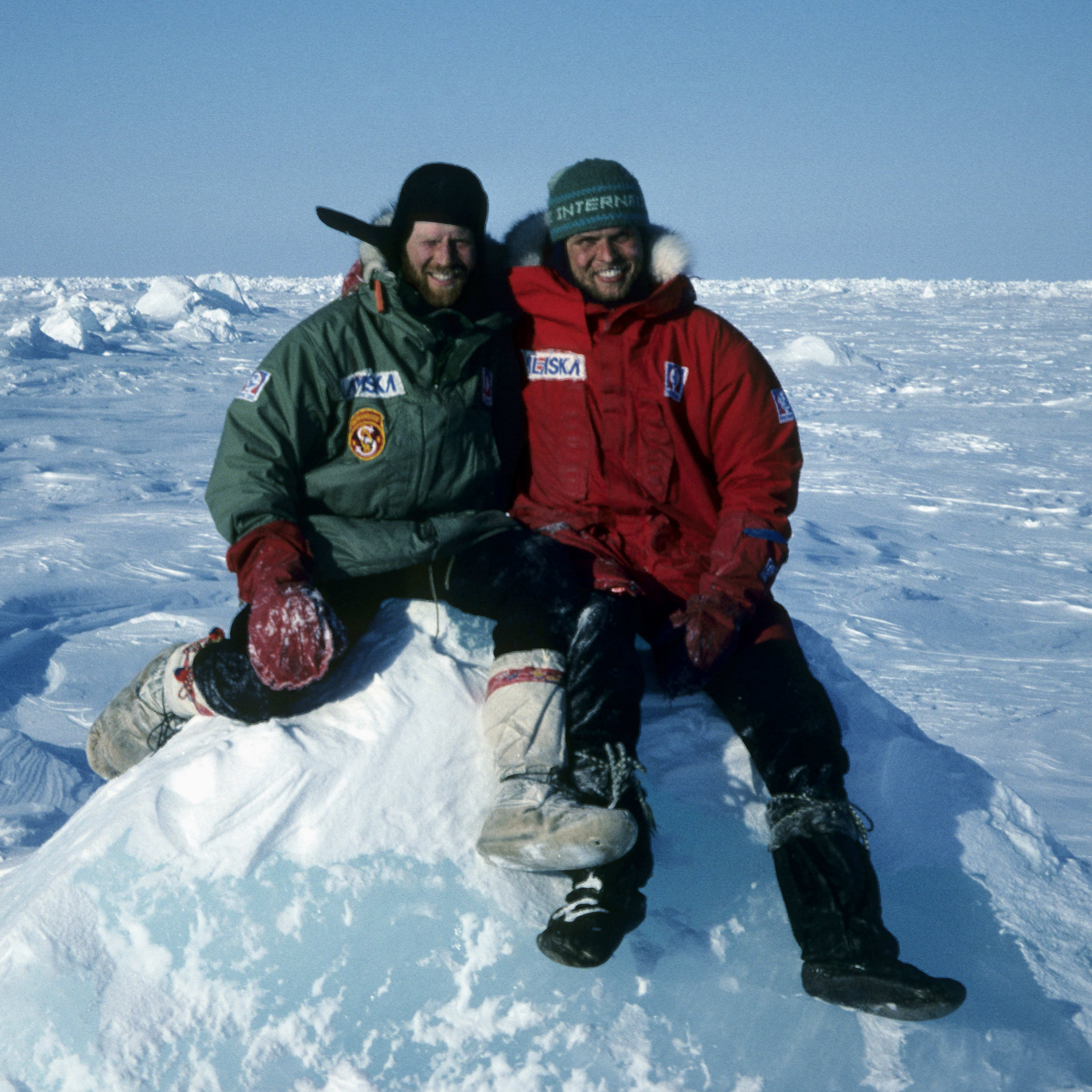 Two men in winter gear sitting on a snowdrift. The man on the left is wearing a green jacket and the man on the right is wearing a red jacket. The background is a snowy, flat landscape.