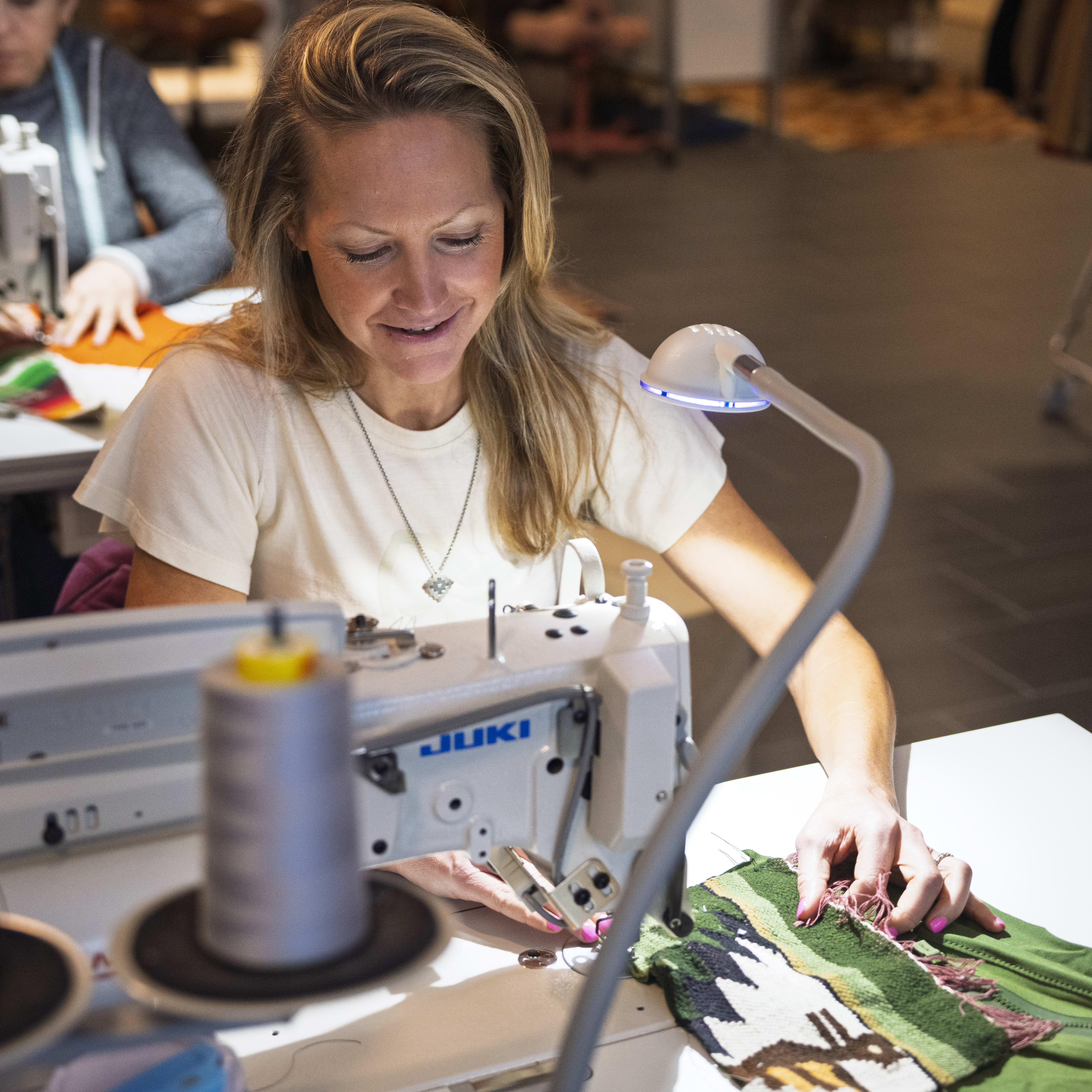 A woman sews a green Norrøna garment at a Juki sewing machine under a bright lamp, smiling. Another seamstress works in the background.
