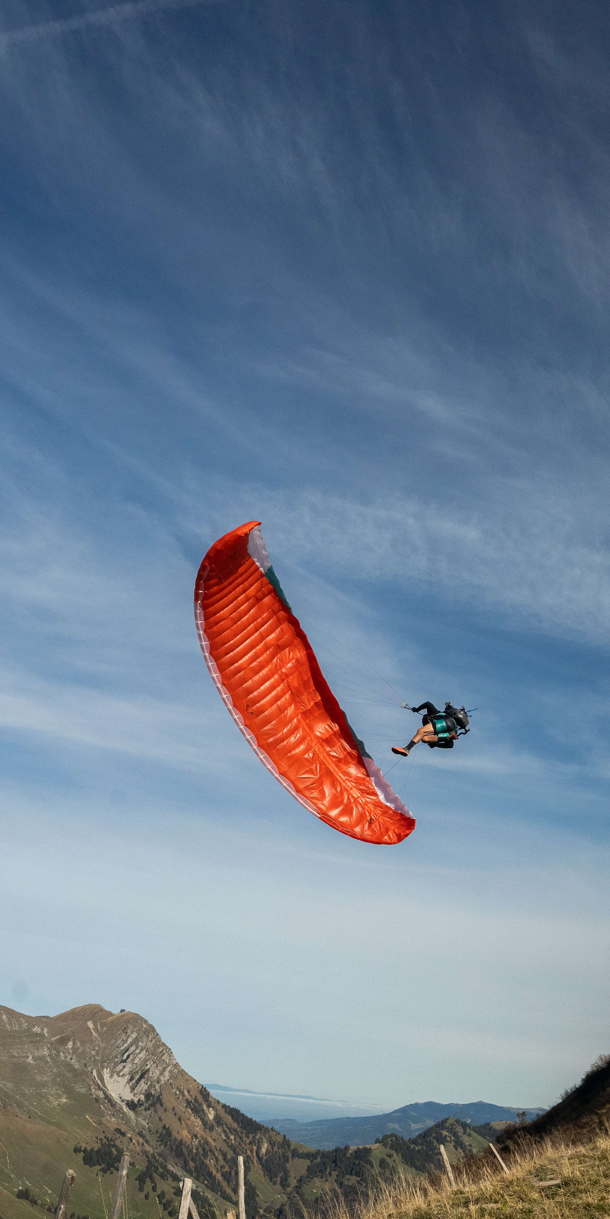 A red paraglider flies over a grassy hillside with a mountain in the background. The sky is blue and clear, with a few wispy clouds.