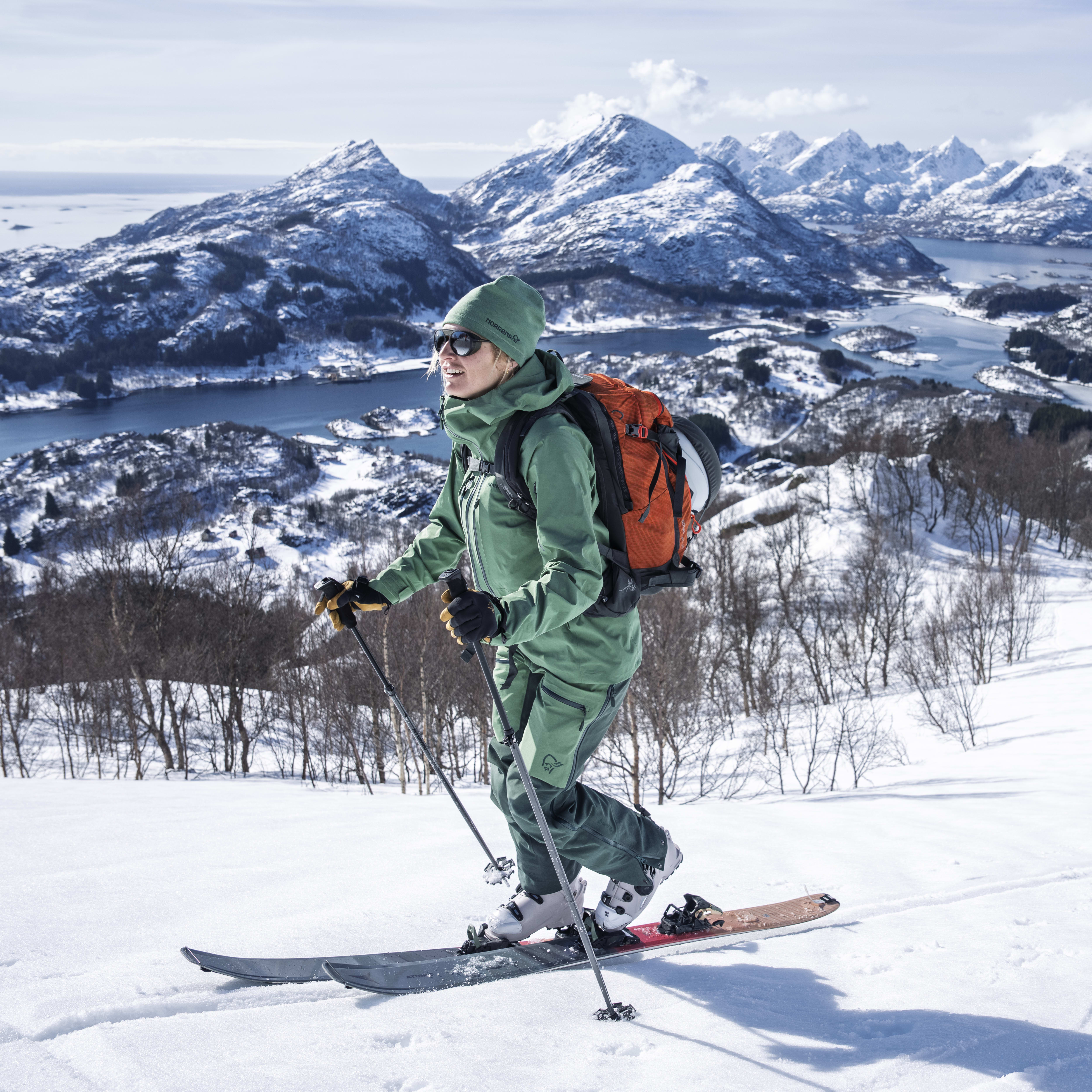 A woman wearing Norrøna gear backcountry skis down a snowy mountain slope with a view of snow capped mountains and a fjord. 
