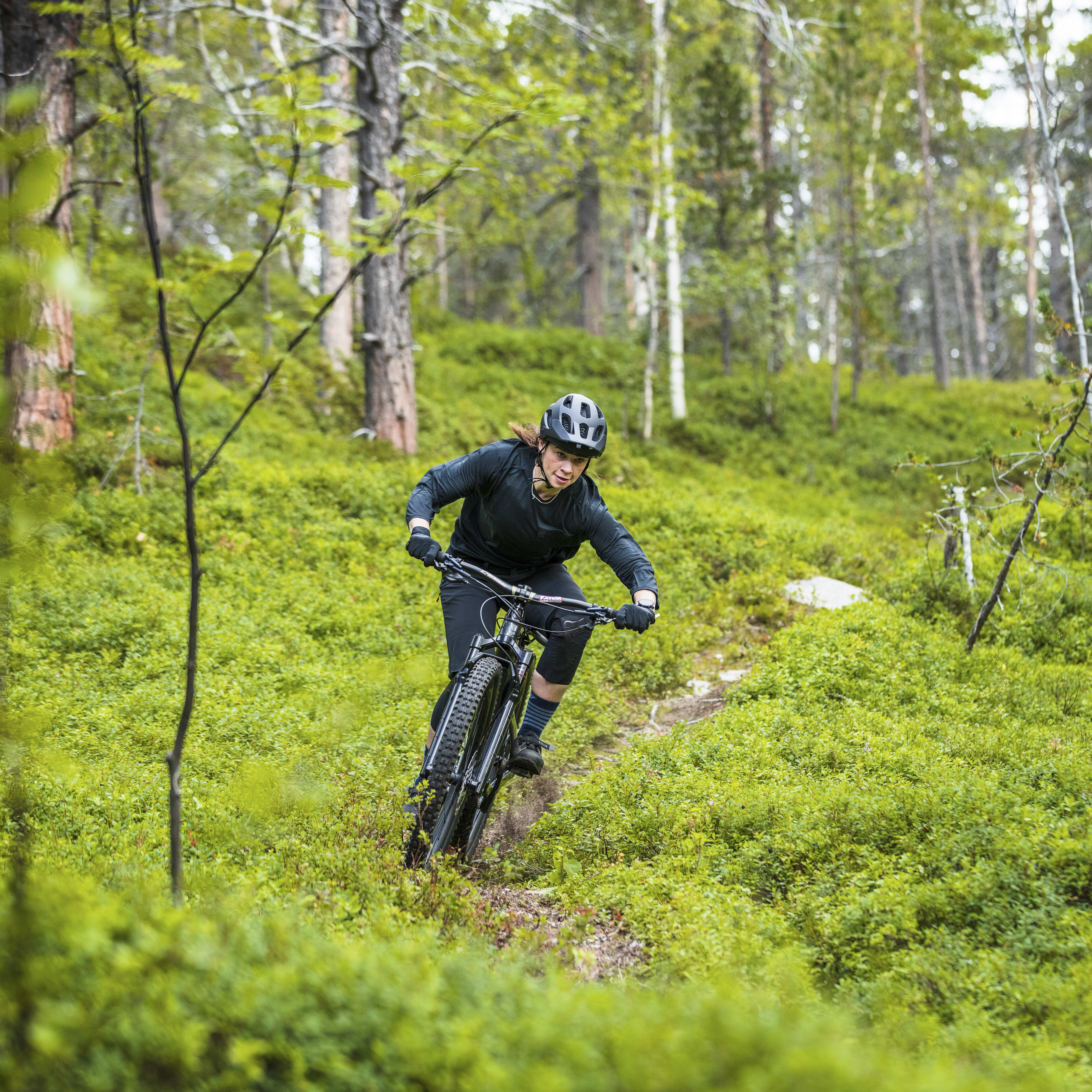 A woman mountain biking through a bright green forest. She wears a helmet and a black Norrøna jacket.