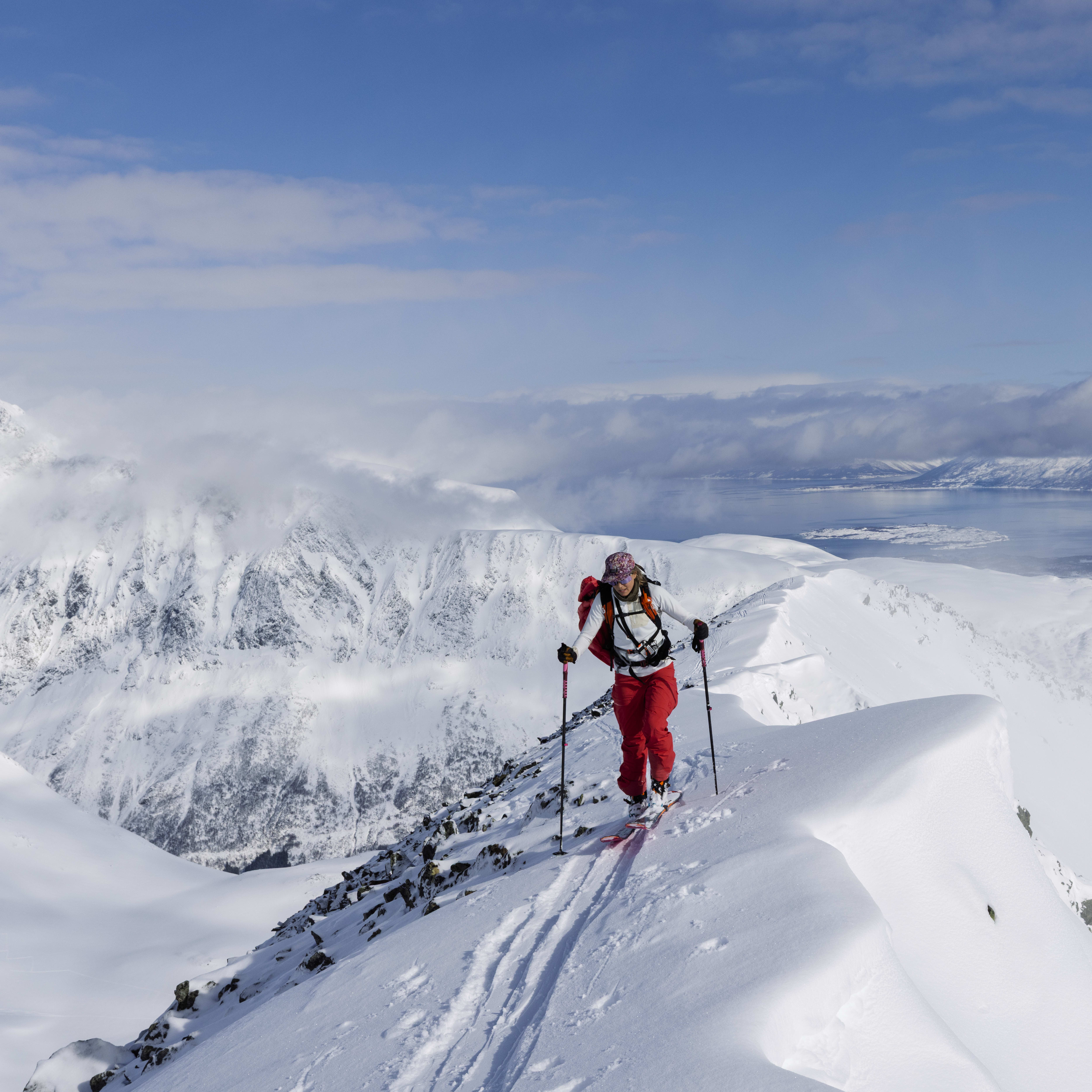 A skier wearing Norrøna red pants ascends a snowy mountain ridge. Mountain peaks and a blue body of water are in the background.
