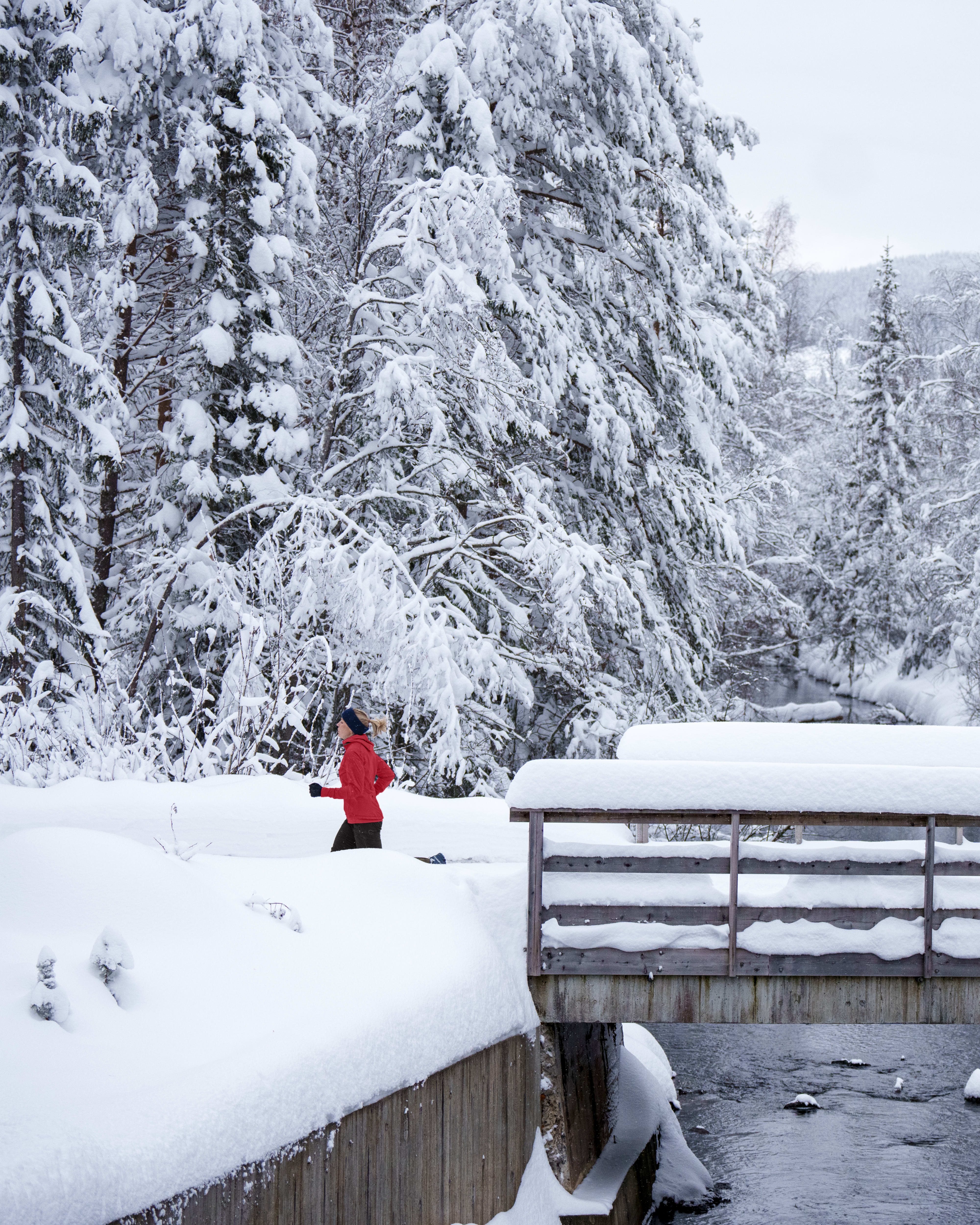 A woman in a red Norrøna jacket runs in a snowy forest next to a bridge over a river. Trees are covered in snow.