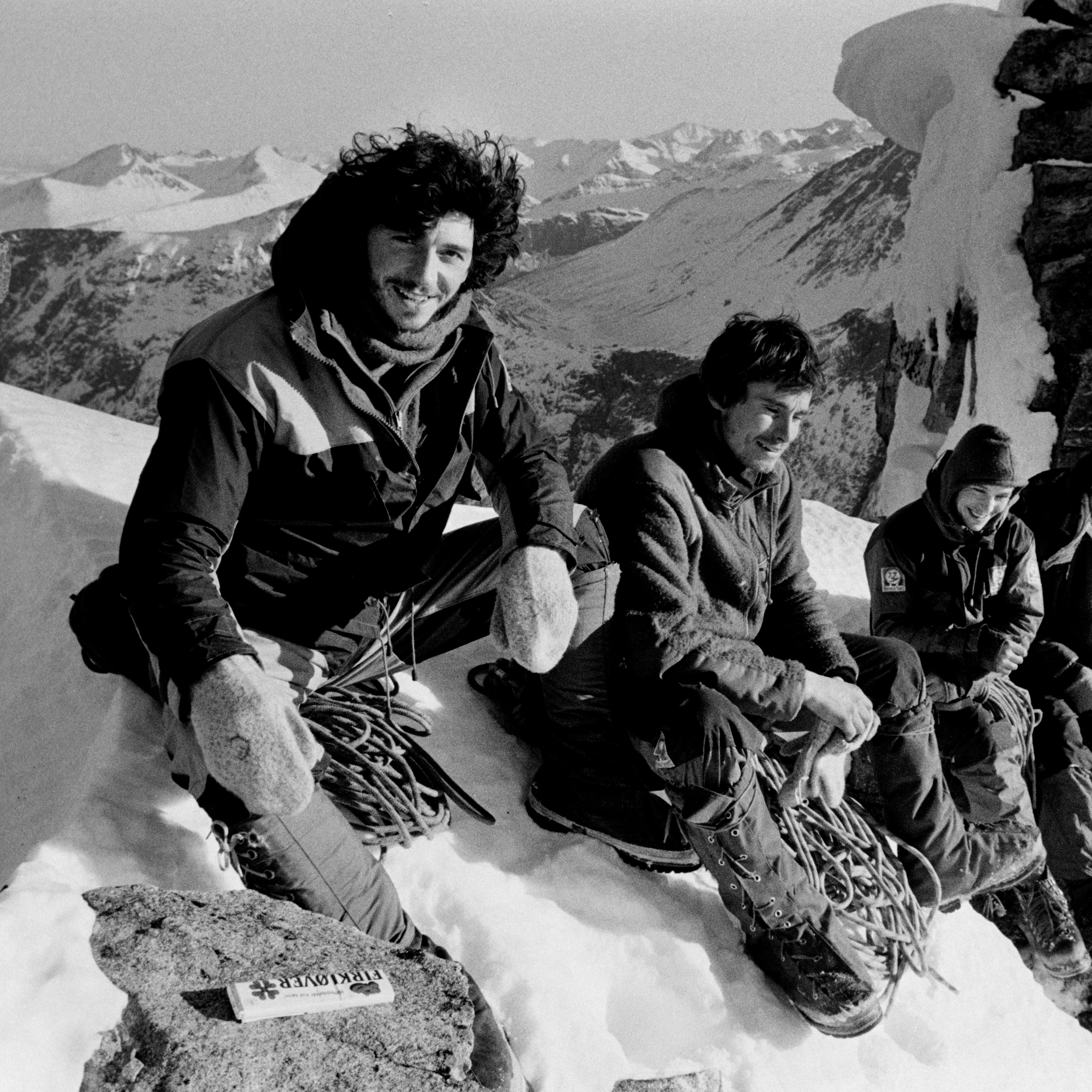 Four climbers are sitting on a snowy mountaintop. They are wearing winter clothing and are surrounded by snow and ice. A coil of rope is beside them. They are all smiling. A mountain range is in the background.