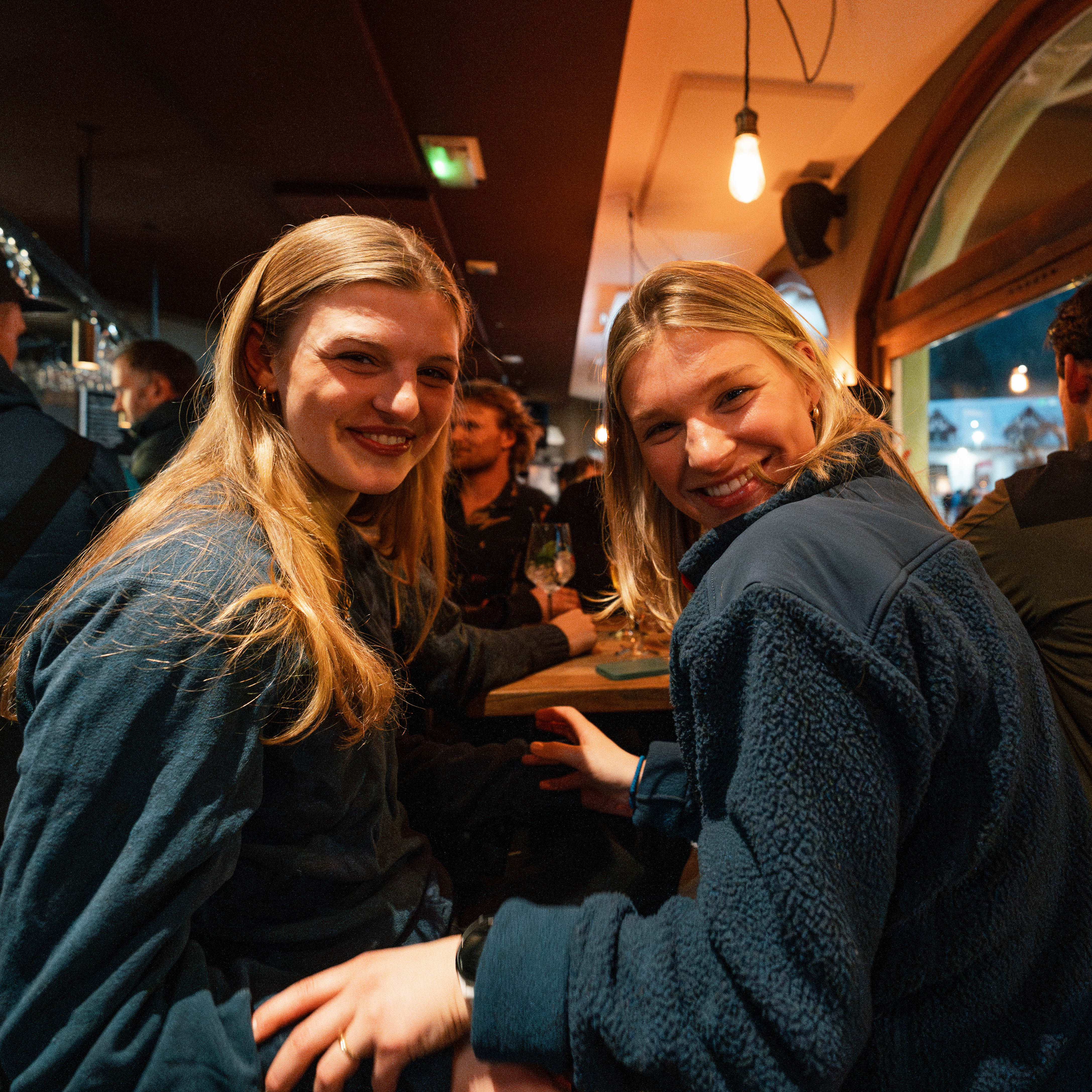Two smiling women in a bar looking at the camera, one wears a blue Norrøna fleece jacket.