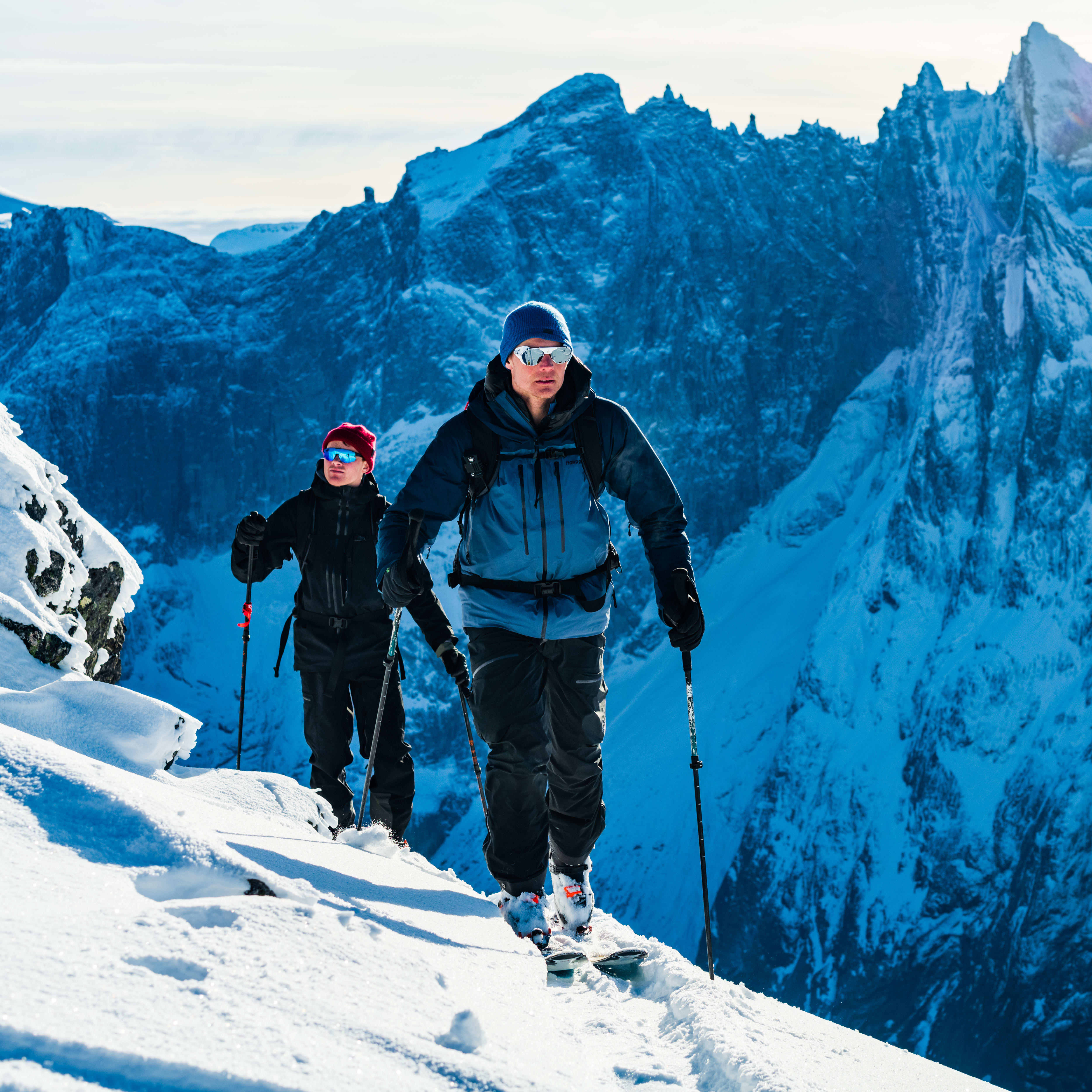 Two people ski down a snow-covered mountainside. They are wearing Norrøna gear and have poles. The mountain is in the background.