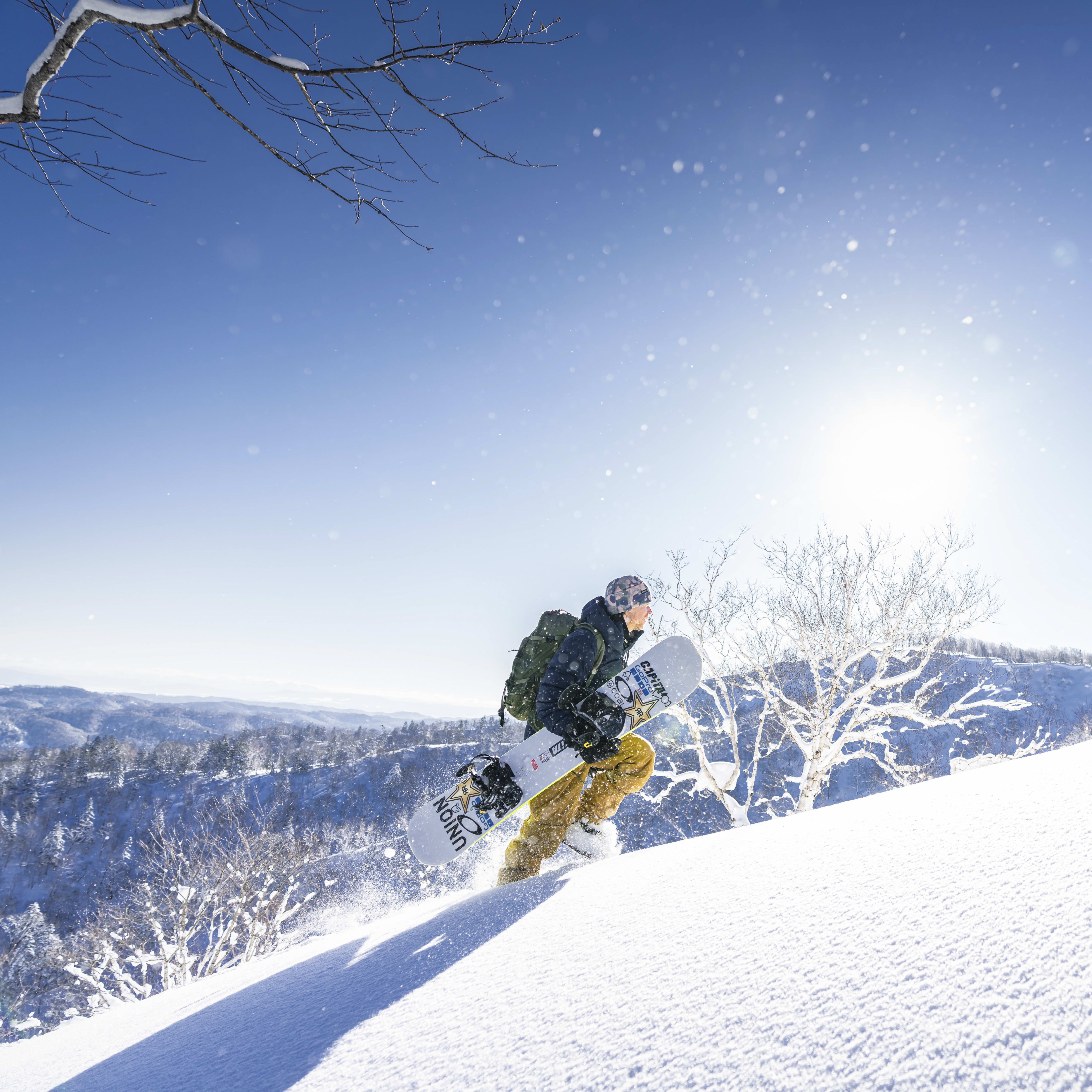 A snowboarder in a Norrøna jacket is carving down a snowy mountain slope. The sky is blue and there are trees in the background.
