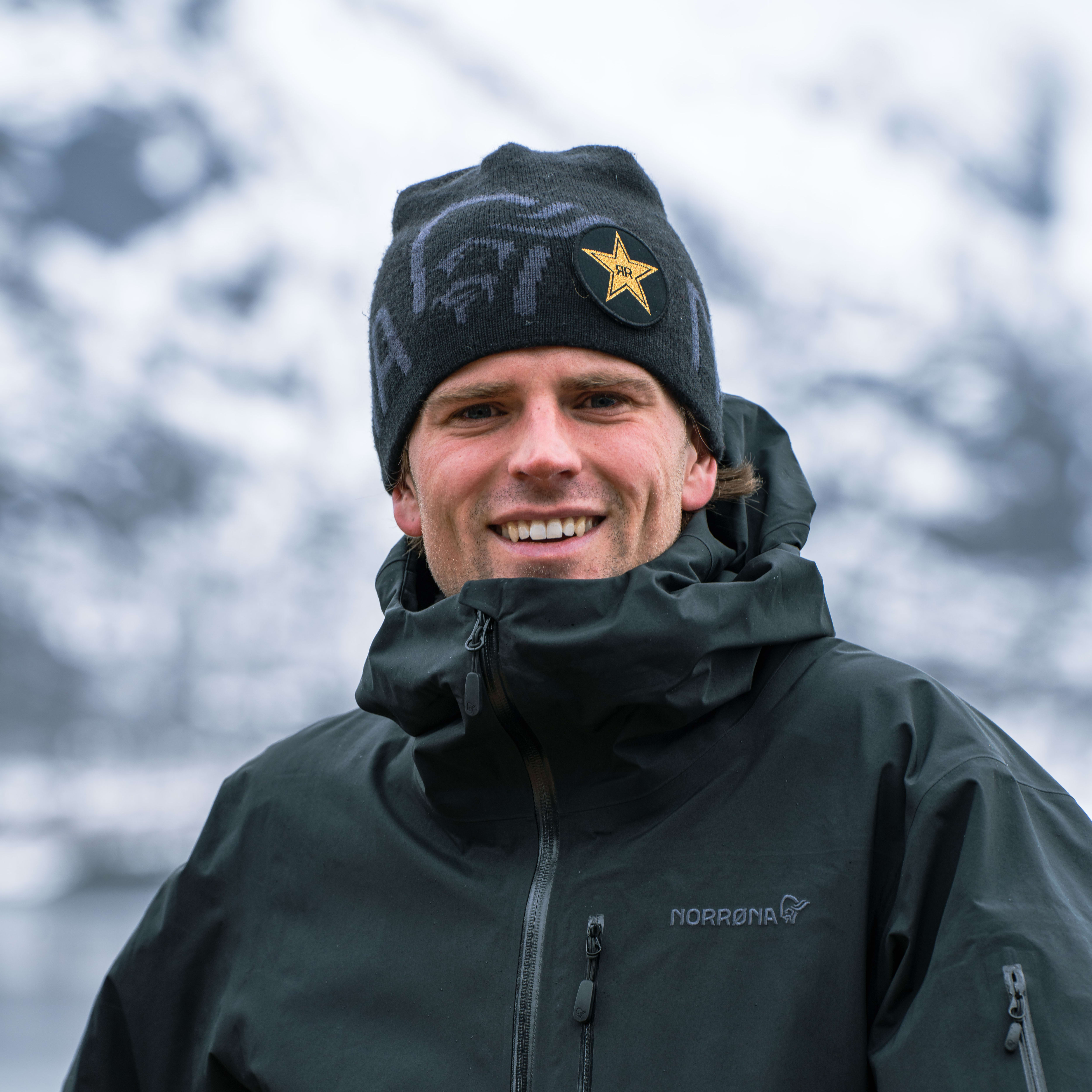 A man wearing a black Norrøna jacket, a black beanie with a yellow star, is smiling. He is standing outside in front of a blurred snowy background. 
