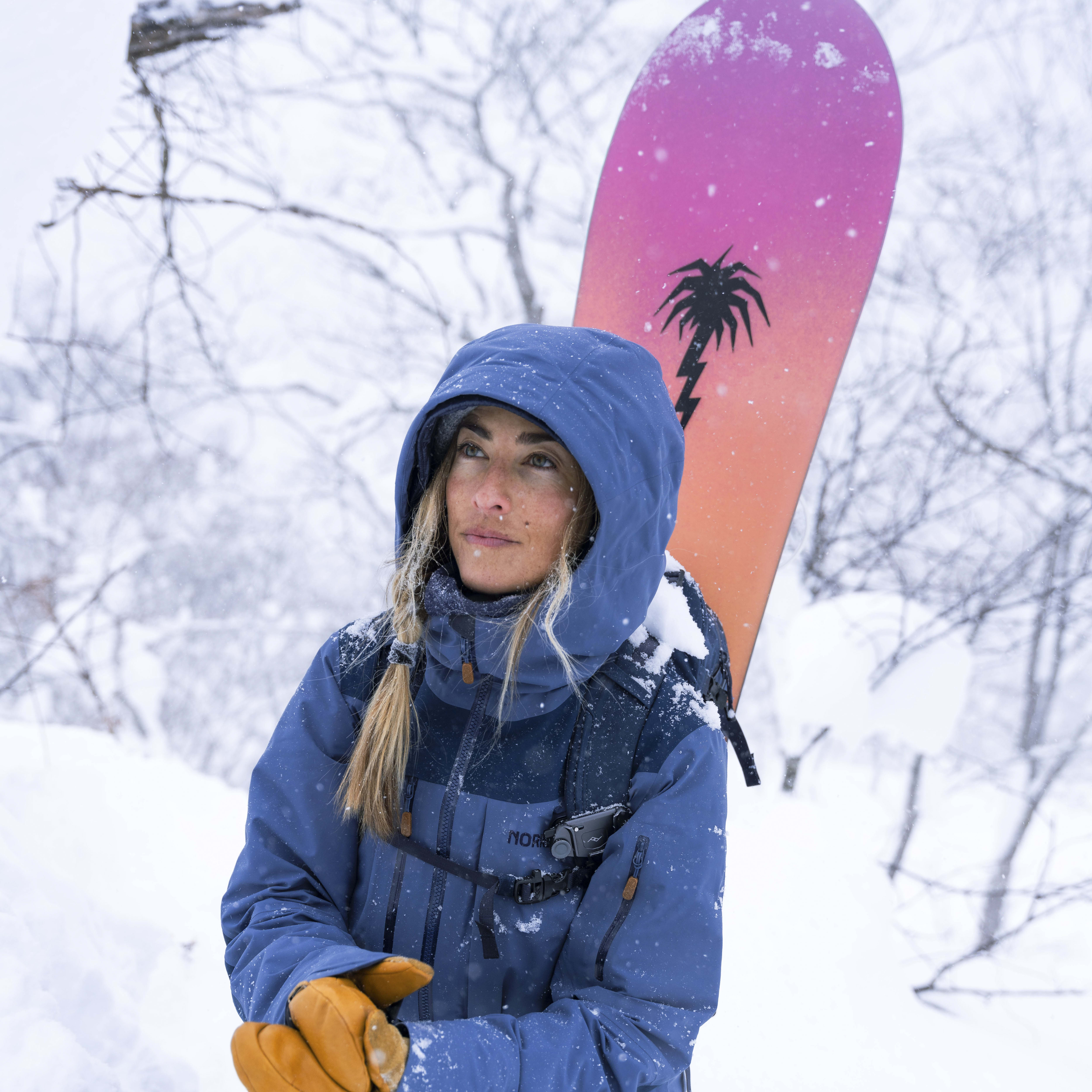 A woman in a blue jacket and brown gloves is kneeling in a snowy field. She is looking upwards, and a snowboard with a pink and orange design is leaning against her back. Snow is falling.