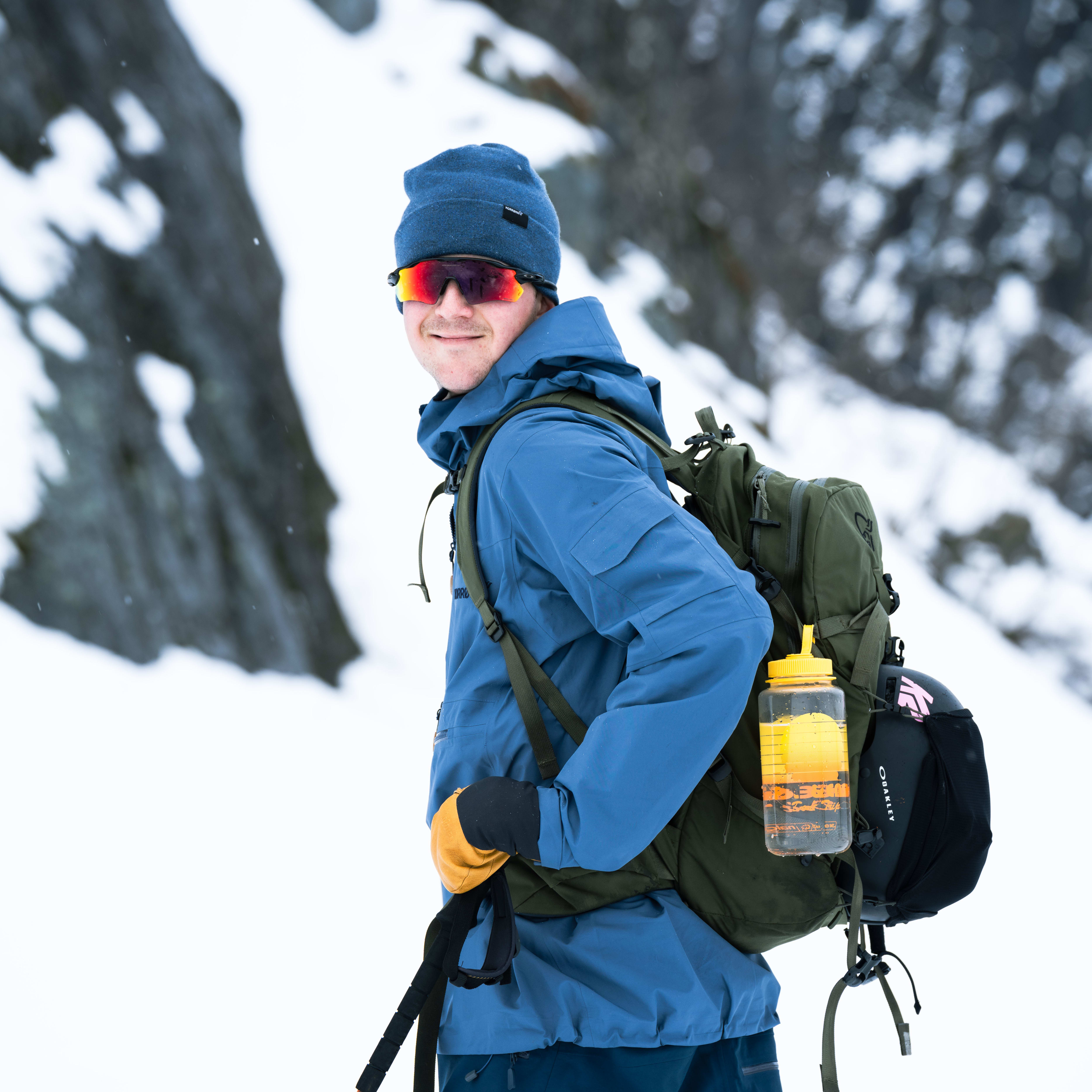 A man wearing a blue Norrøna jacket, blue Norrøna pants, a blue Norrøna beanie, and sunglasses is standing in the snow. He is holding a ski pole and carrying a green backpack. The backpack has a yellow water bottle attached to it. The man is smiling and looking at the camera. He is wearing a black helmet with an Oakley logo. There is snow falling in the background.