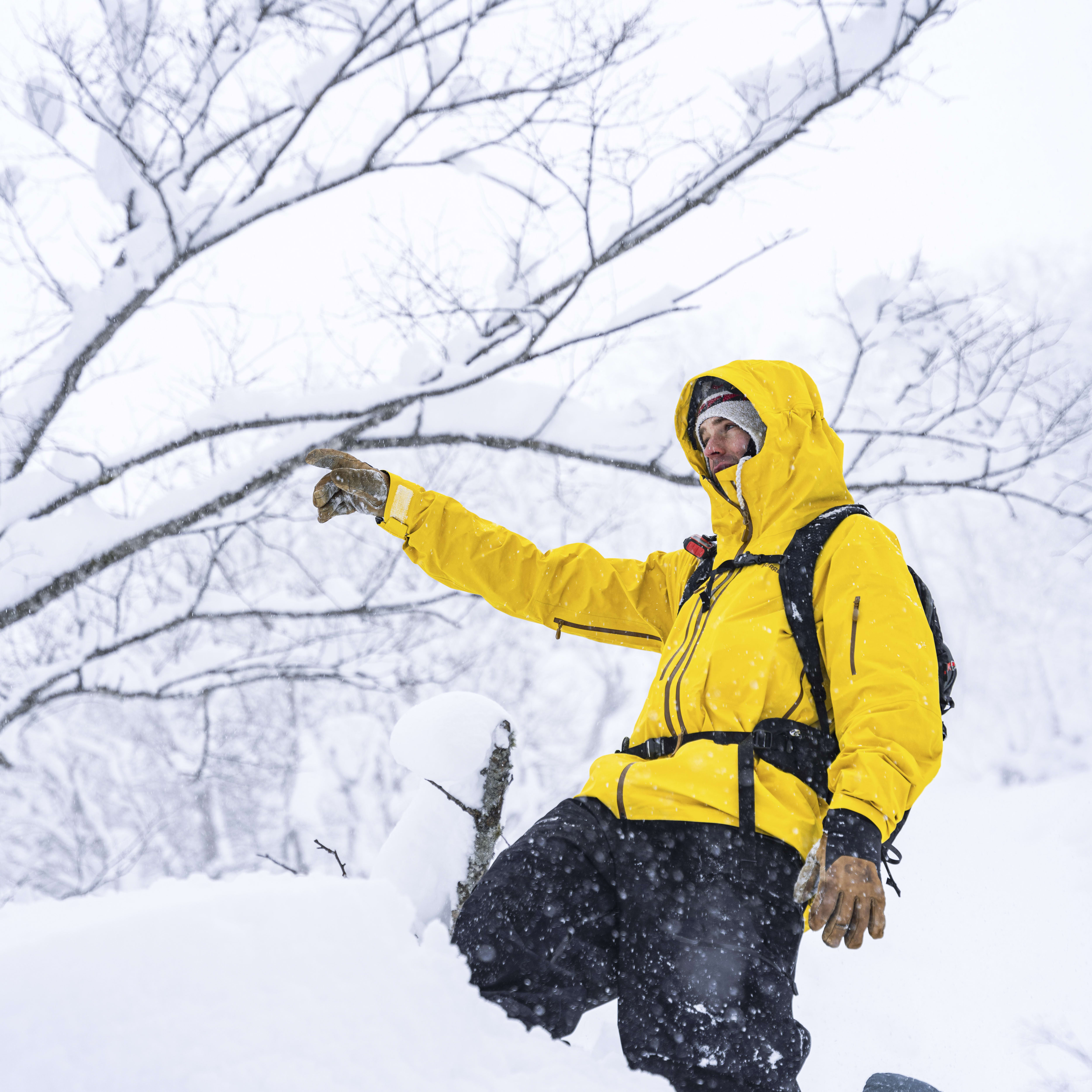 A person in a yellow jacket and black pants is partially hidden in a snowdrift. The person is pointing with their right hand and snow is falling.